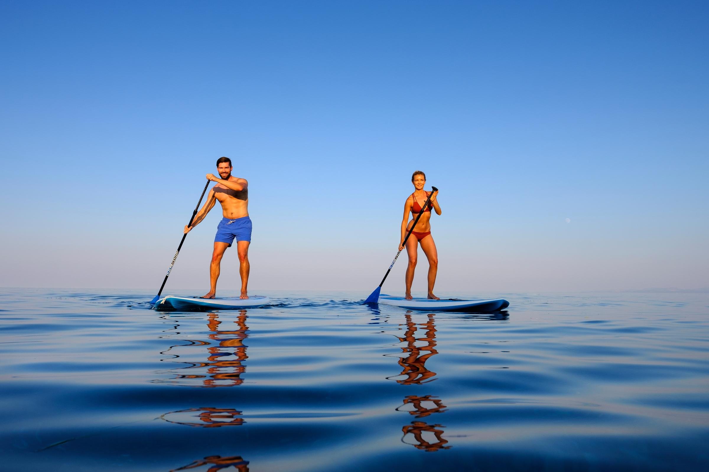 Zwei Personen beim Stand-Up-Paddling auf ruhigem Wasser, Sonnenlicht, blauer Himmel.