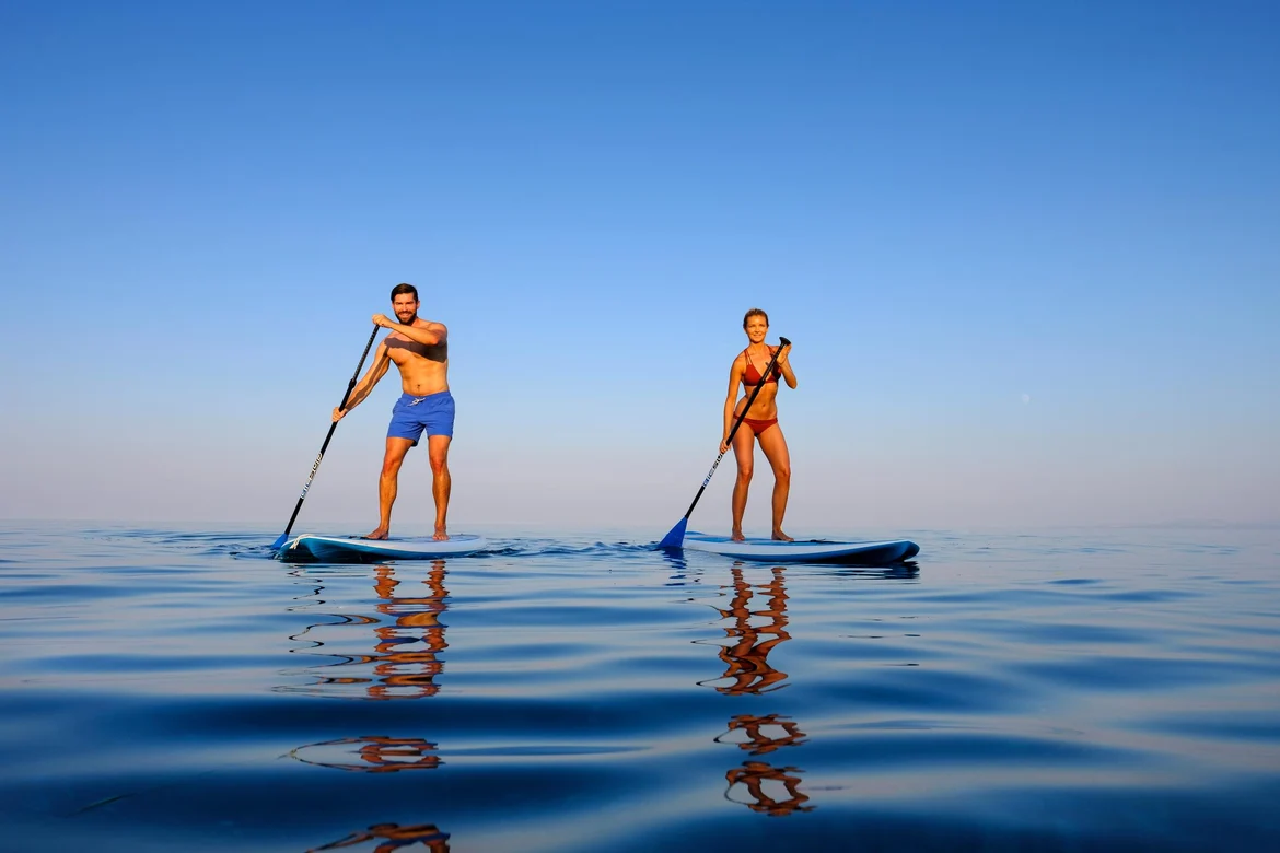 Zwei Personen beim Stand-Up-Paddling auf ruhigem Wasser, Sonnenlicht, blauer Himmel.