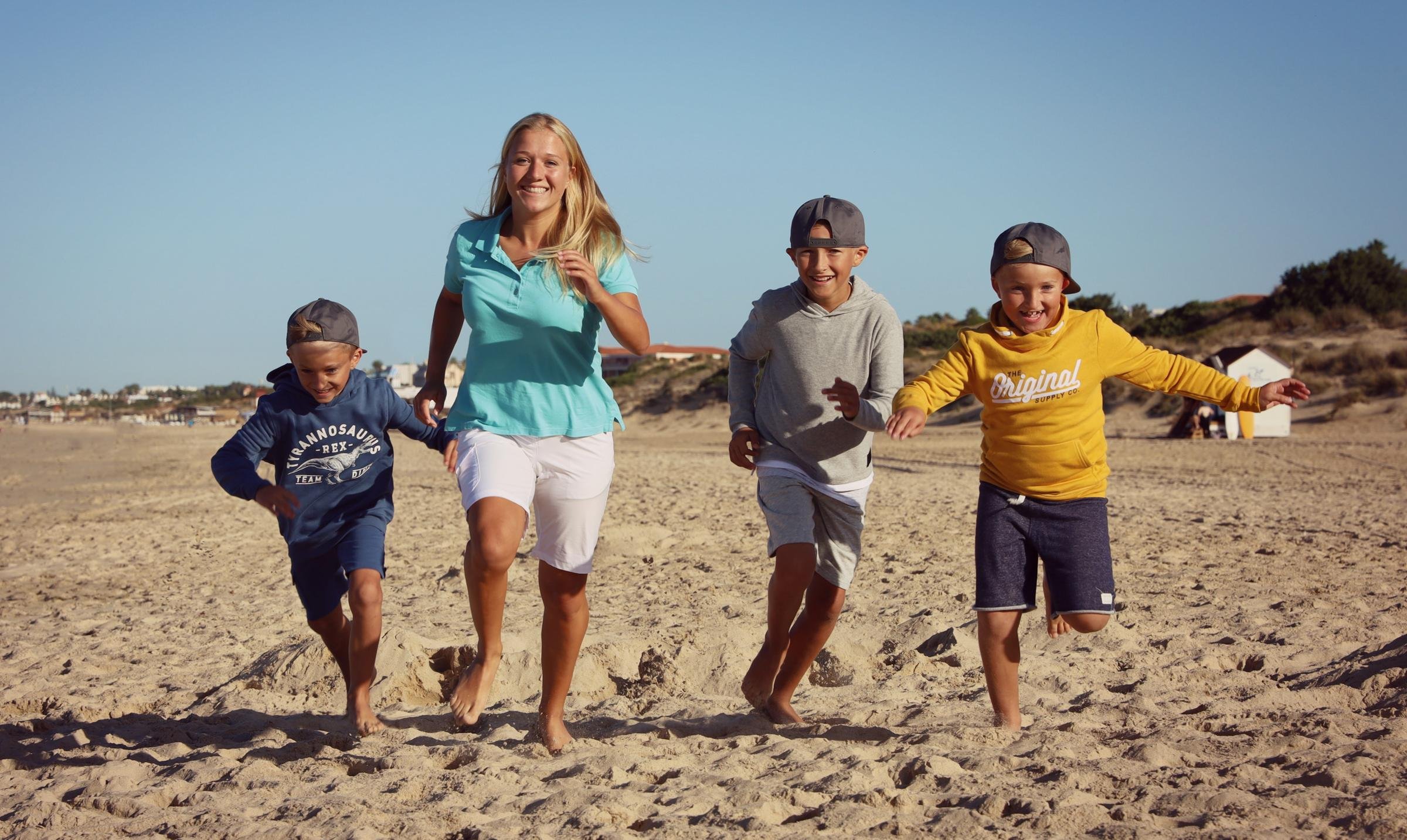 Eine Frau mit drei Kindern läuft barfuß am Strand entlang, Sonne und Meer im Hintergrund