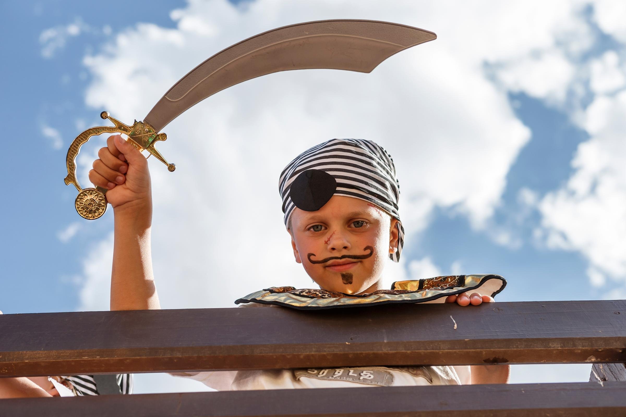 Ein Kind in Piratenkostüm mit Spielschwert auf einem Surfbrett im Himmel
