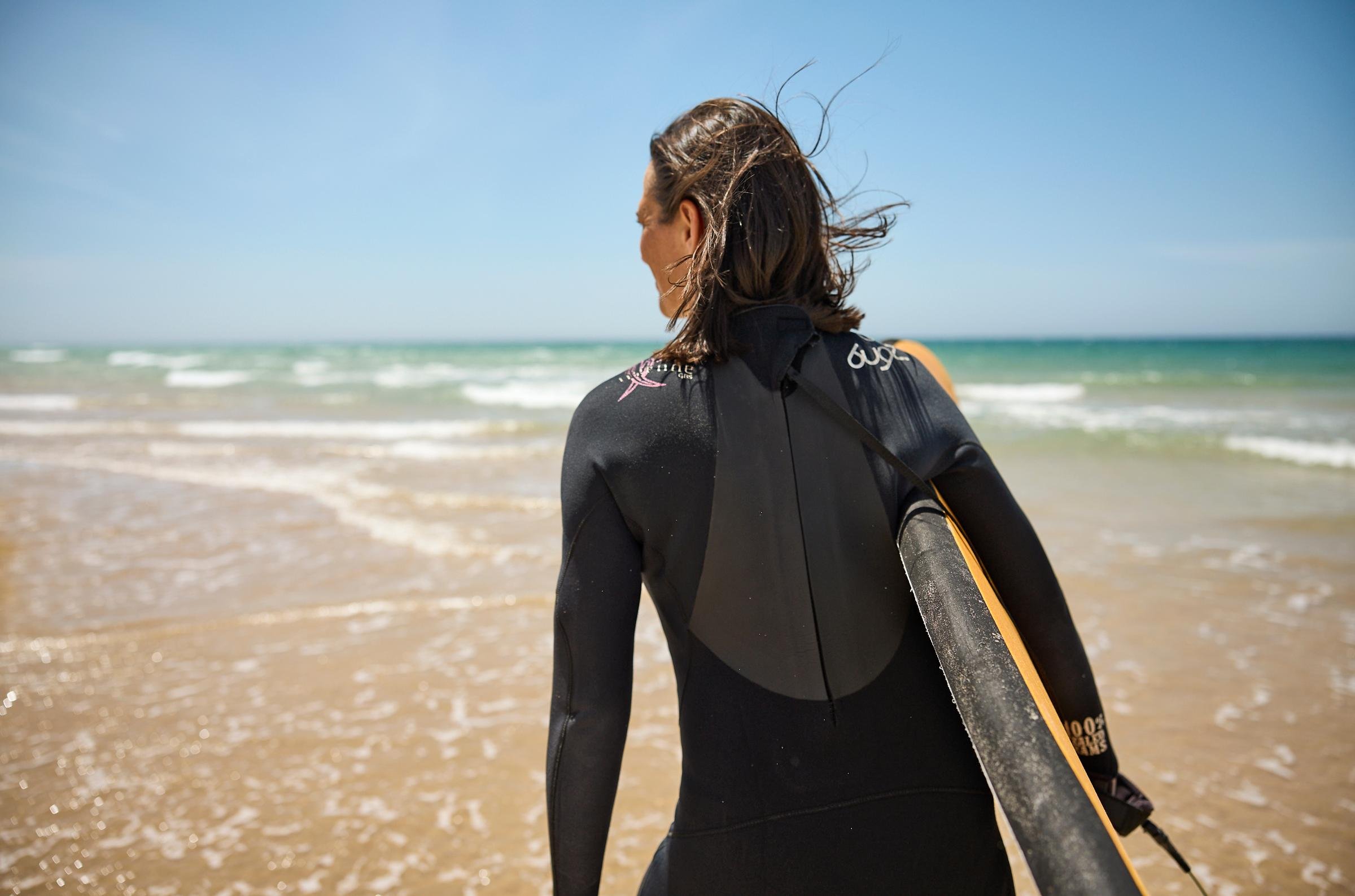 Frau mit Surfausrüstung am Strand bei sonnigem Himmel, Premium Cluburlaub bei Aldiana
