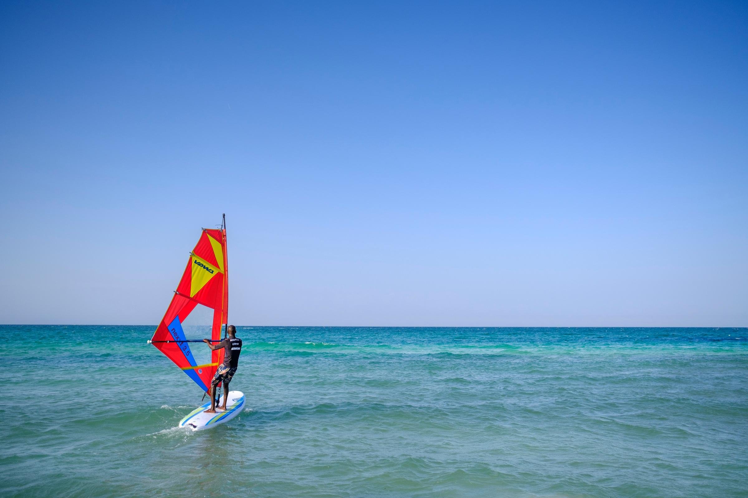 Ein Mann windet auf einem Surfbrett mit roter Segel auf dem Meer, blauer Himmel