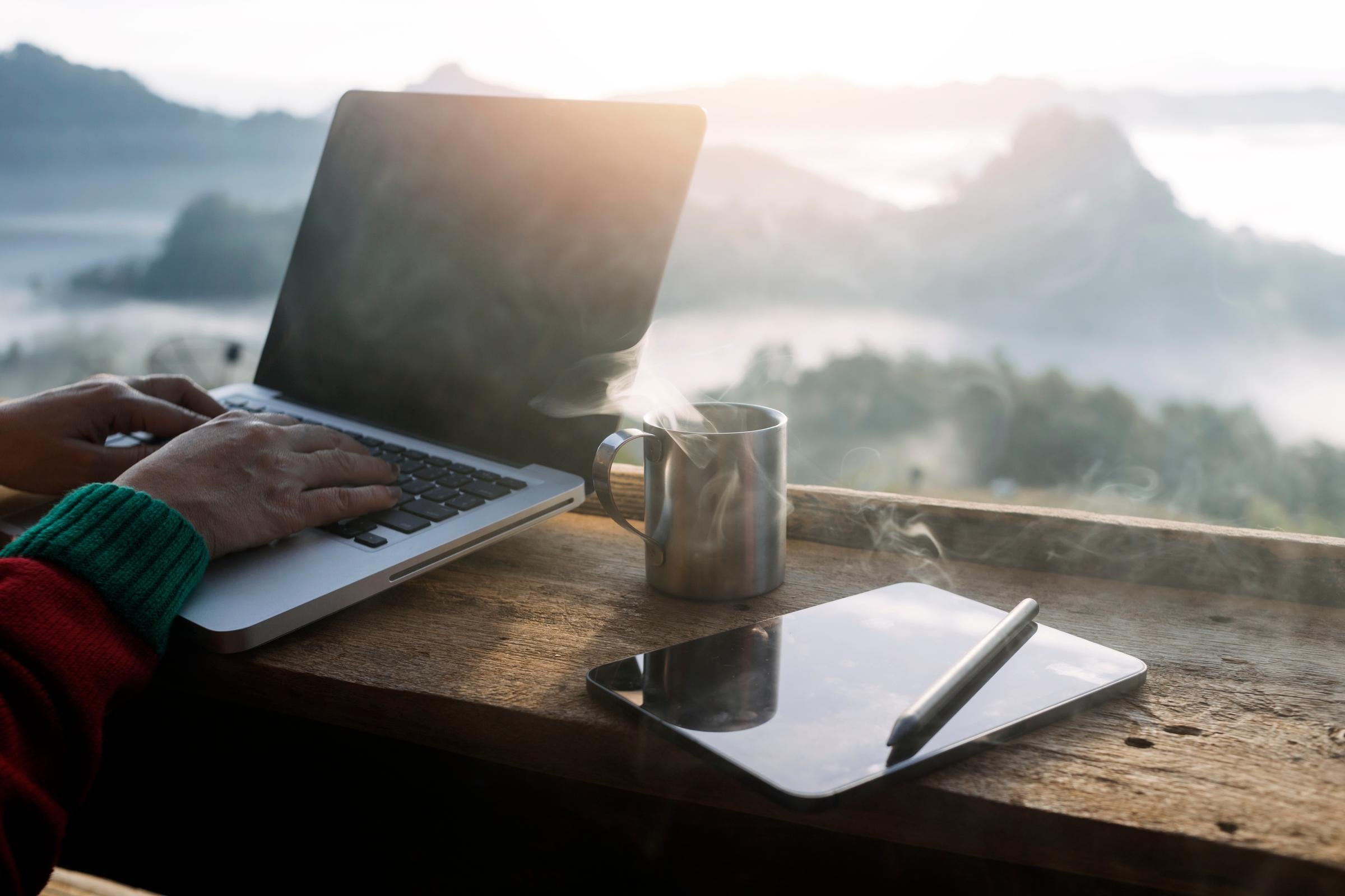 Person arbeitet an Laptop mit Kaffee und Tablet in schönem natürlichen Urlaubsambiente
