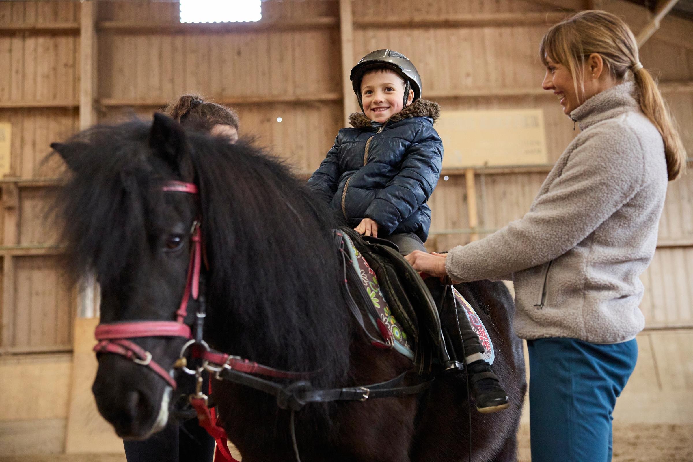 Mädchen reitet auf einem Pony im Innenraum eines Reitstalls, betreut von einer Erwachsenen