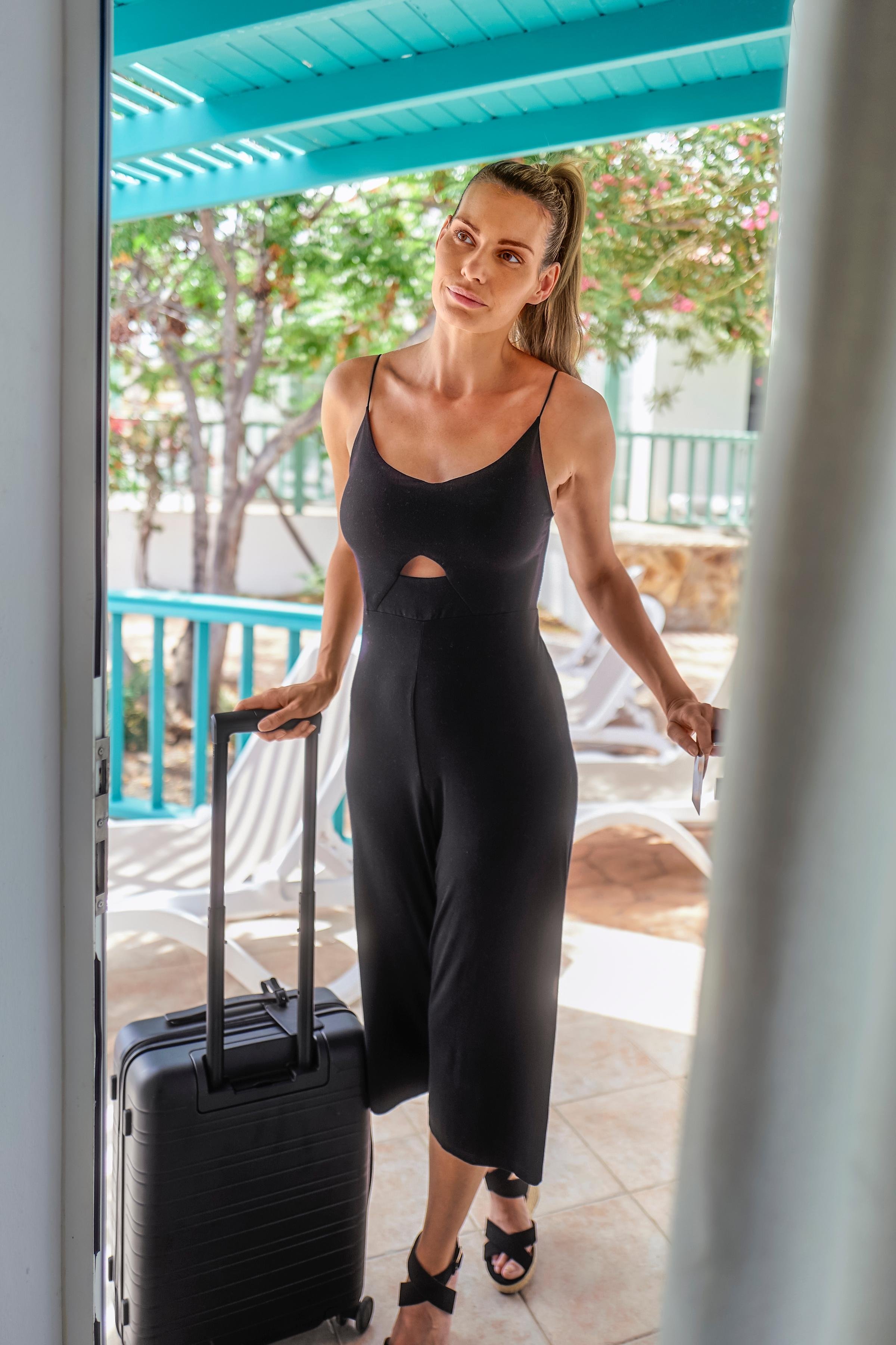 Frau mit einem schwarzen, eleganten Jumpsuit und Gepäck auf sonniger Terrasse bei Aldiana