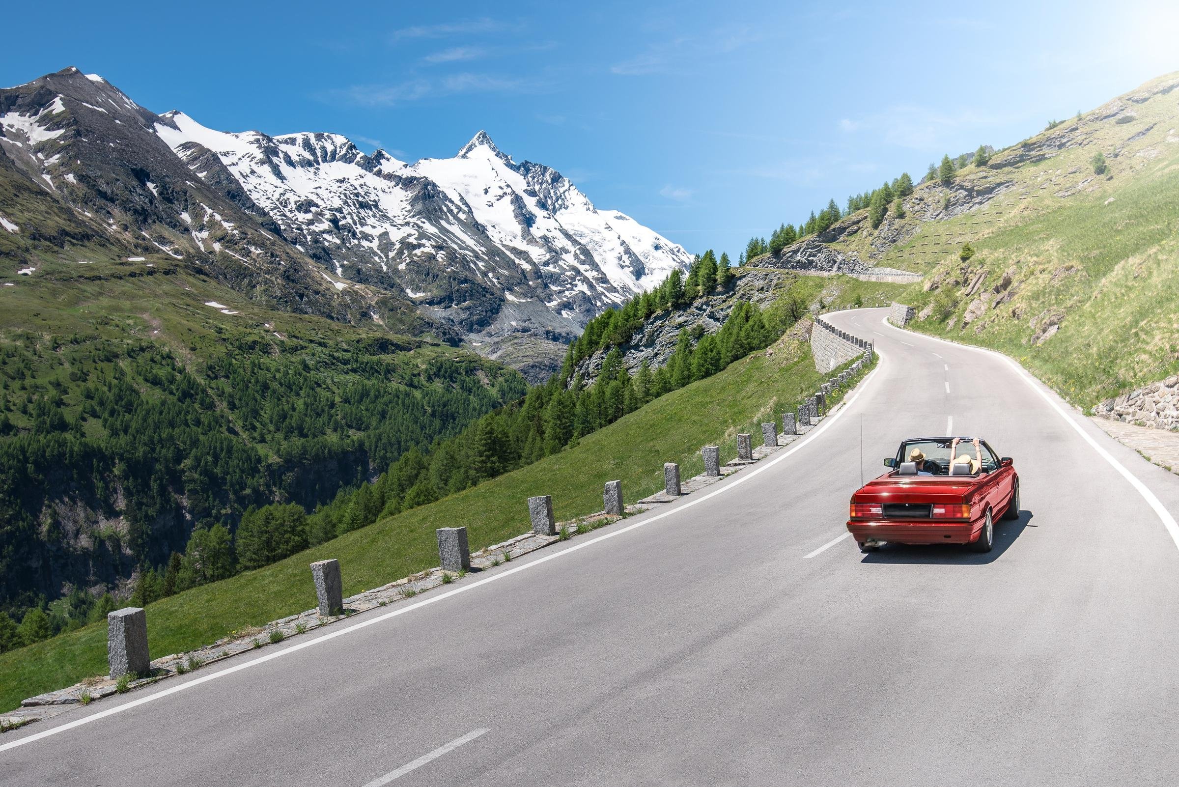 Ein rotes Cabrio fährt auf einer kurvigen Bergstraße mit Blick auf schneebedeckte Berge bei sonnigem Himmel