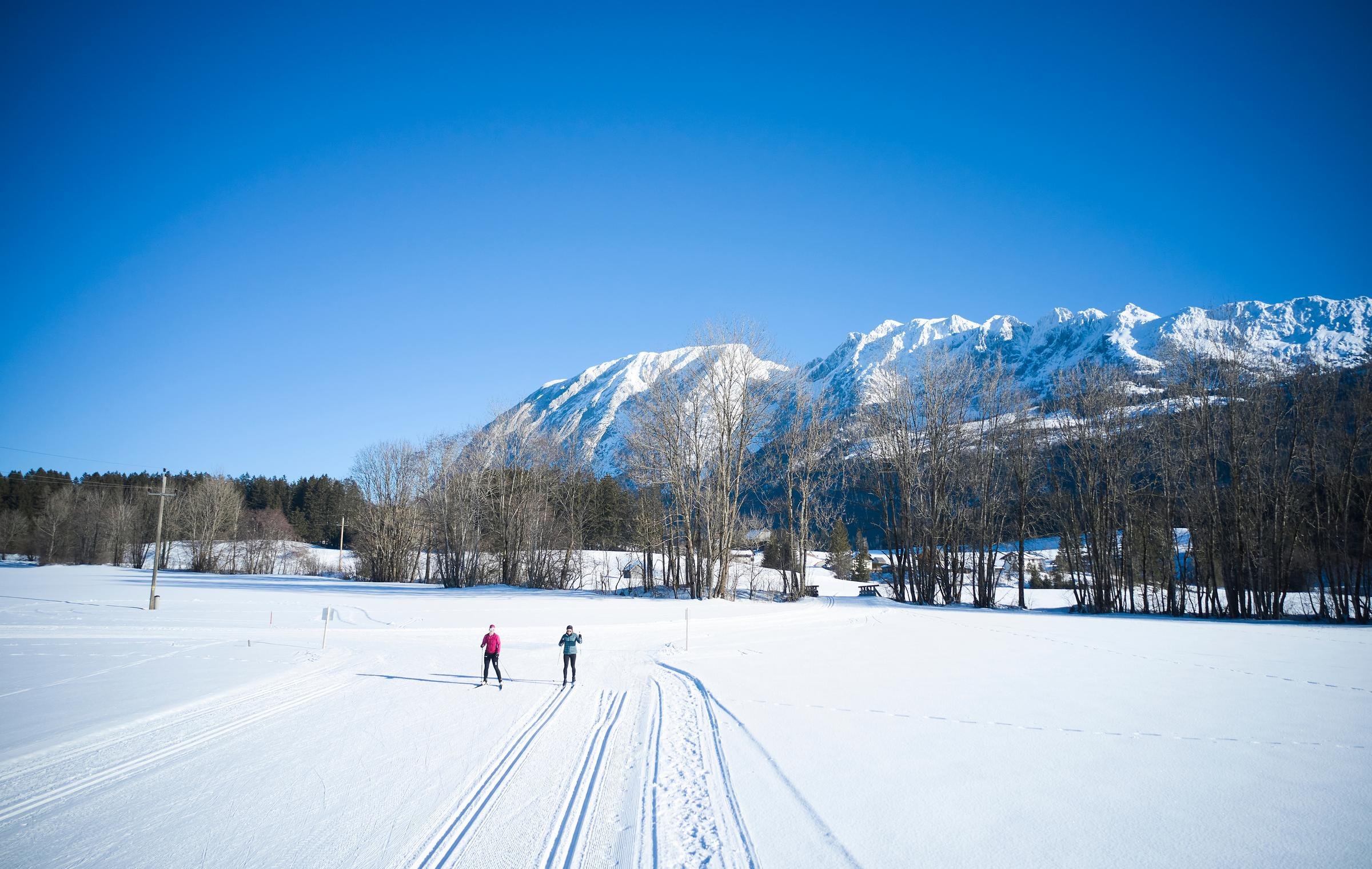 Zwei Skifahrer auf verschneitem Feld vor Bergen, klarer Himmel, winterliche Landschaft