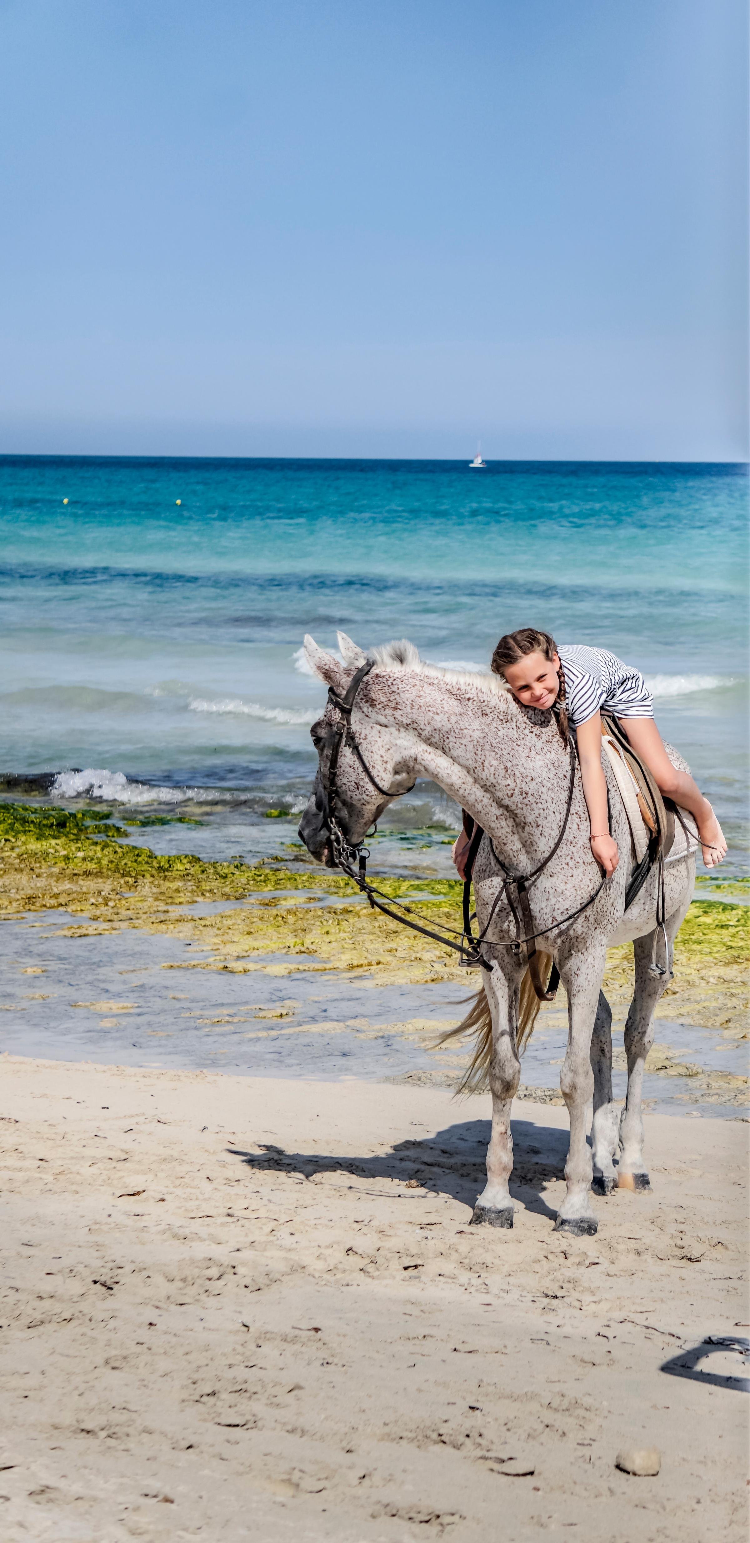 Kind people reiten auf einer weißen Pferdefigur am Strand bei Aldiana, blaues Meer im Hintergrund