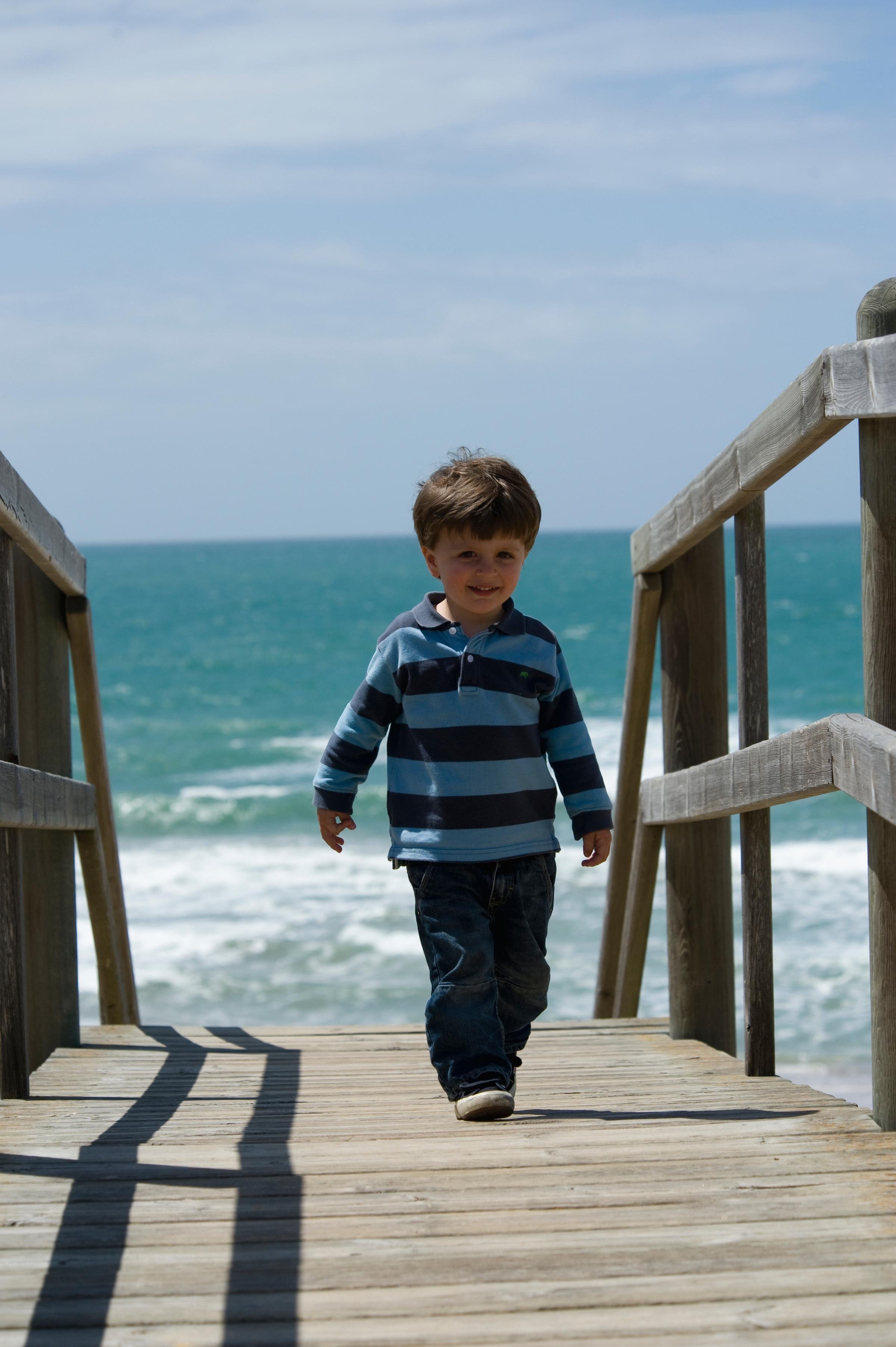 Junge im striped Shirt läuft auf Holzsteg zum Strand mit Meer und Himmel im Hintergrund