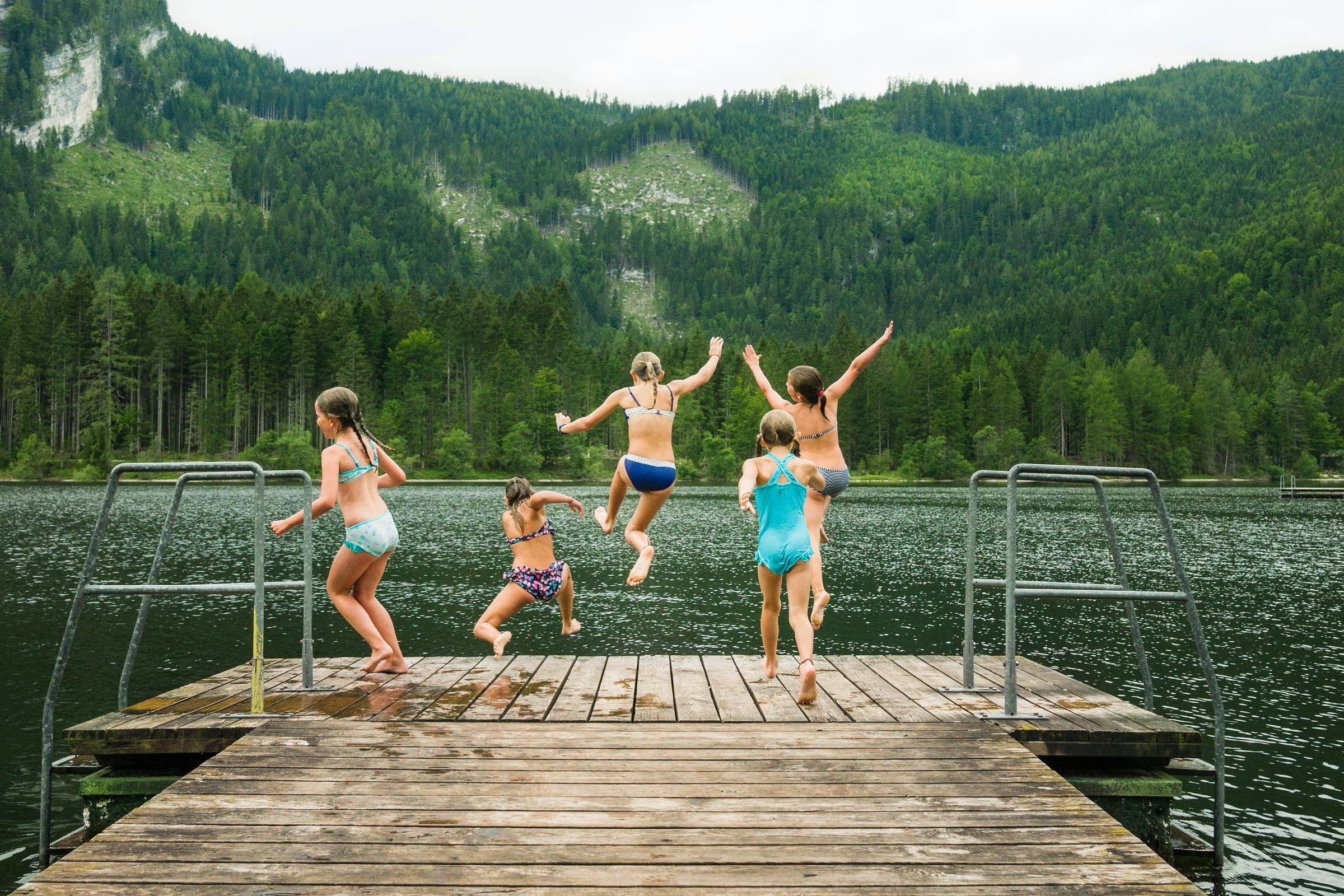 Vier Kinder springen fröhlich vom Steg ins klare Wasser in einer natürlichen Seenlandschaft