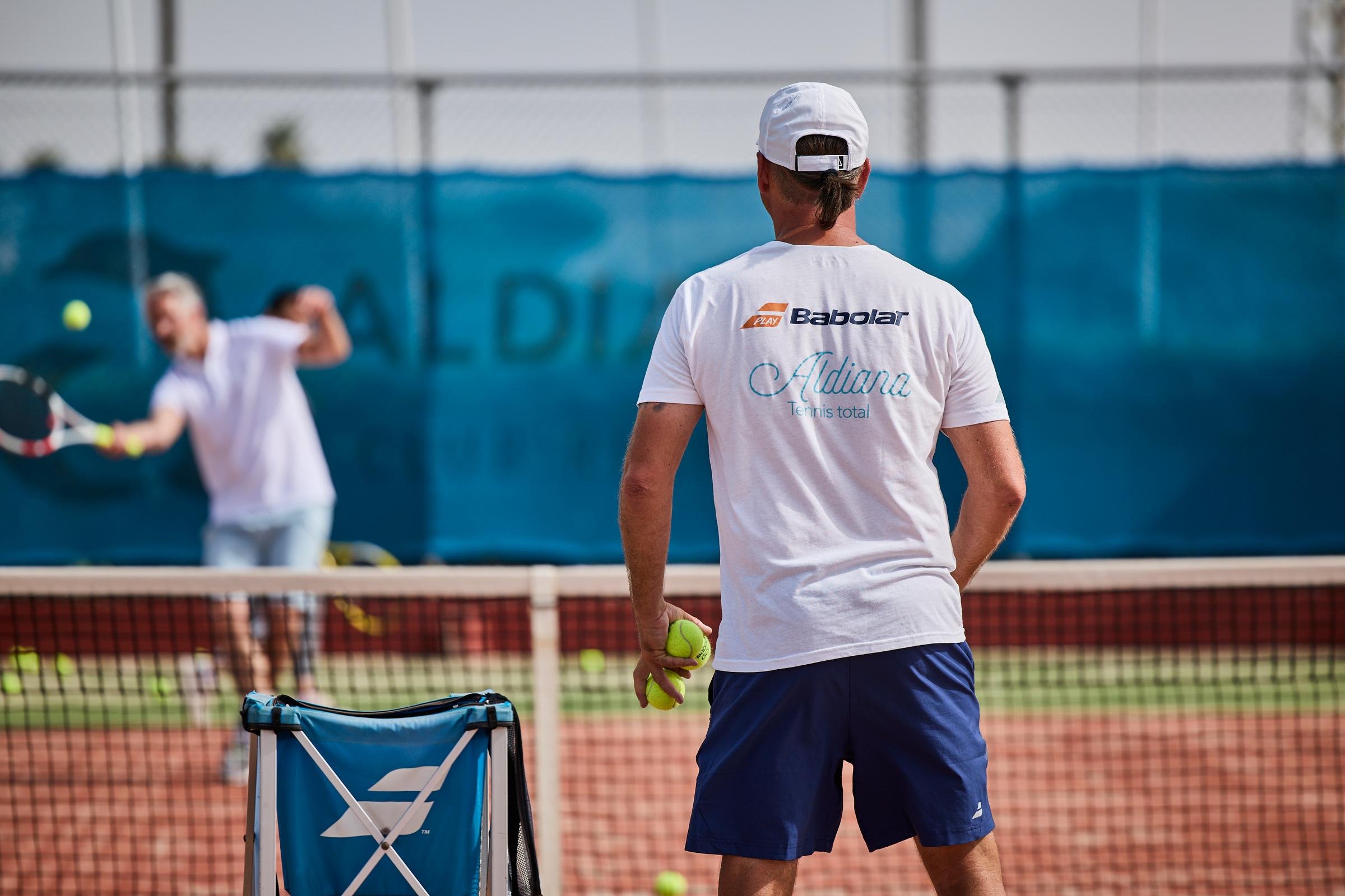 Ein Mann spielt Tennis auf einem Außenplatz mit Blick auf ein Netz und eine blaue Windschutzscheibe