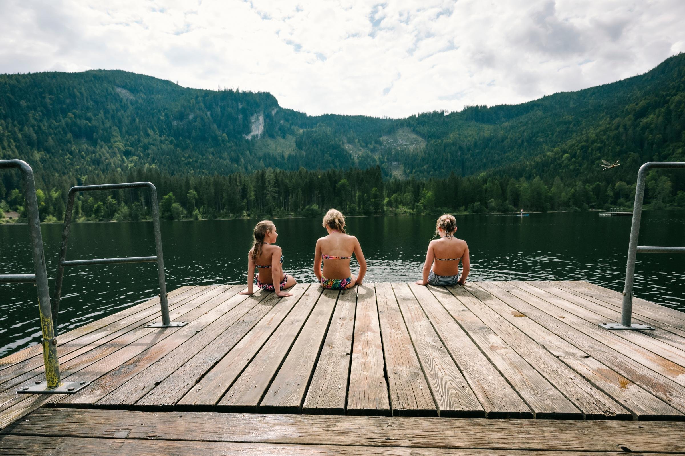 Drei Kinder sitzen auf einem Steg am ruhigen See mit Bergen im Hintergrund, entspannt und idyllisch