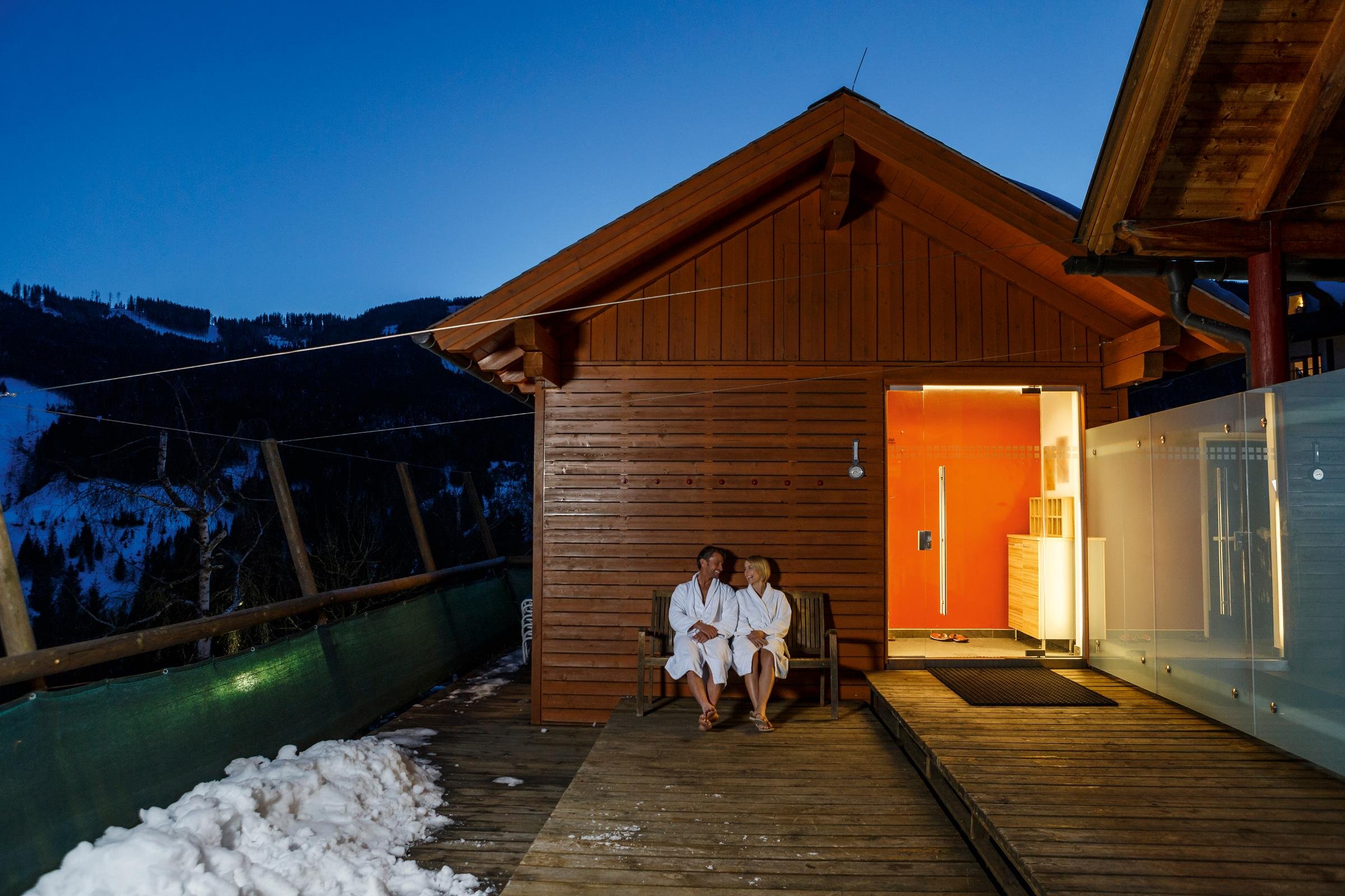 Zwei Personen im Bademantel sitzen auf einer Terrasse vor einem Holzhaus bei Sonnenuntergang