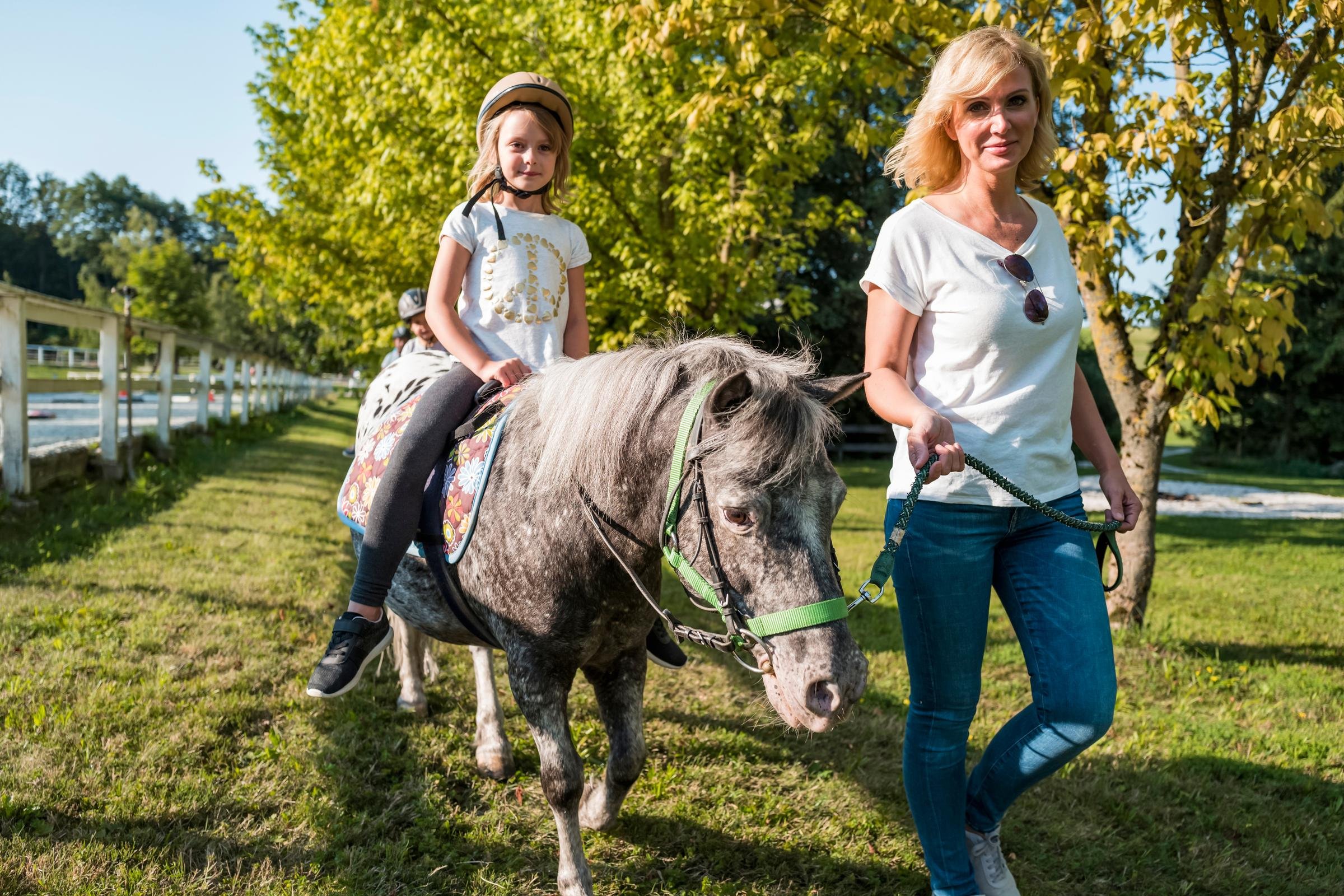 Frau führt Pony bei sonnigem Ausflug im Grünen, Kind reitet auf Pferd, idyllisches Umfeld