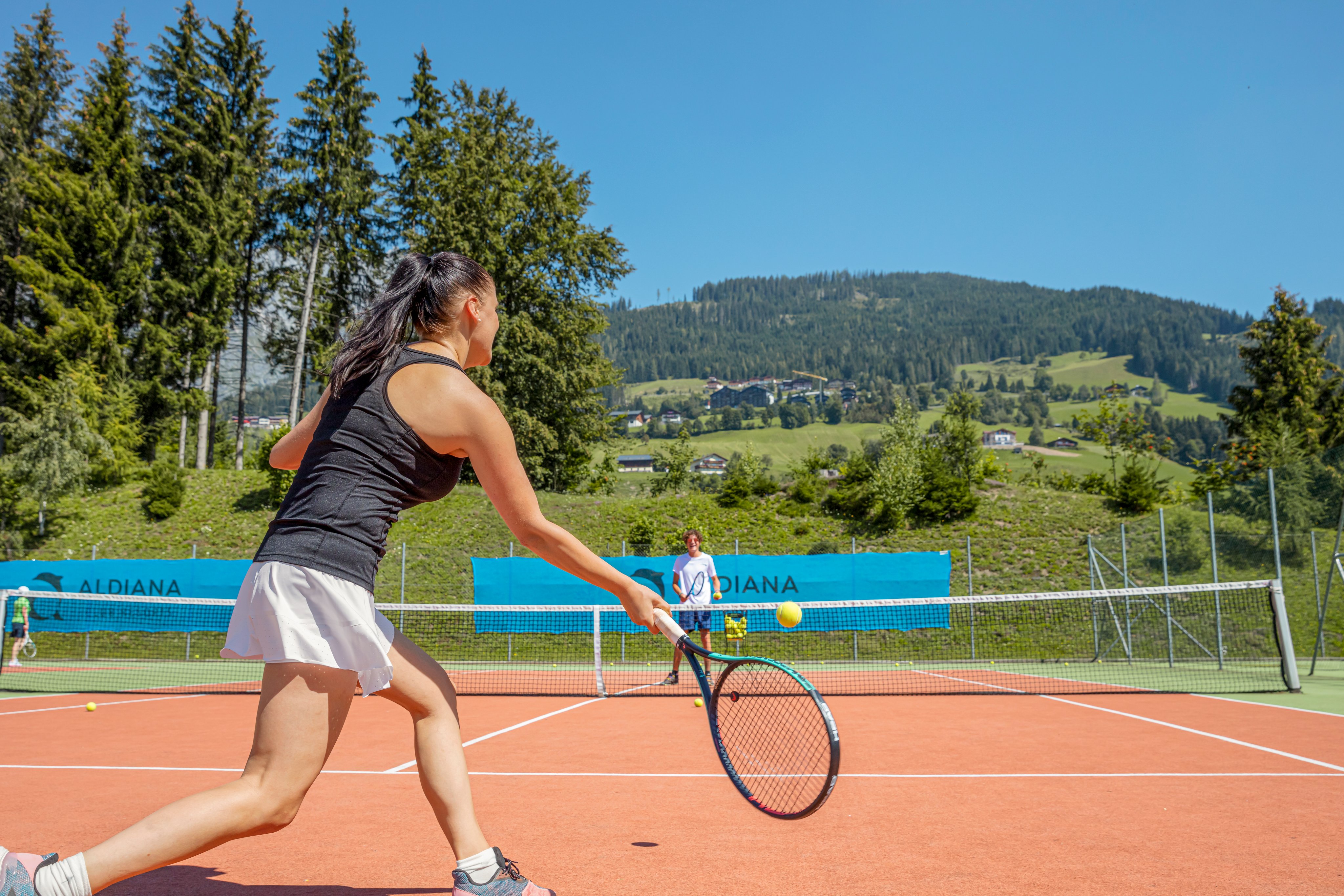 Eine junge Frau spielt Tennis auf einem roten Platz vor einer grünen Berglandschaft bei Aldiana