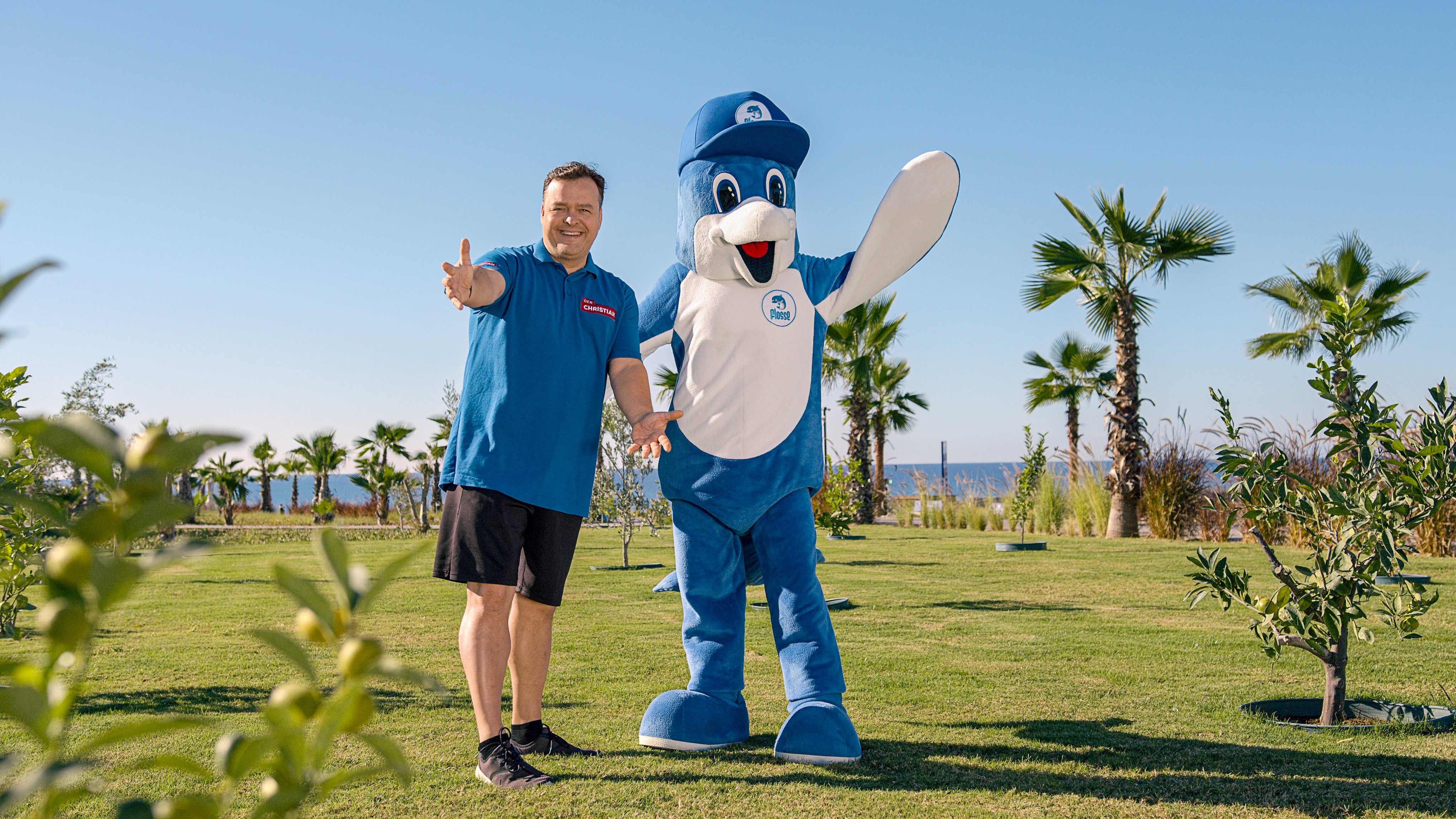 Ein Mensch mit einem Maskottchen in einem gepflegten Garten mit Palmen im Hintergrund