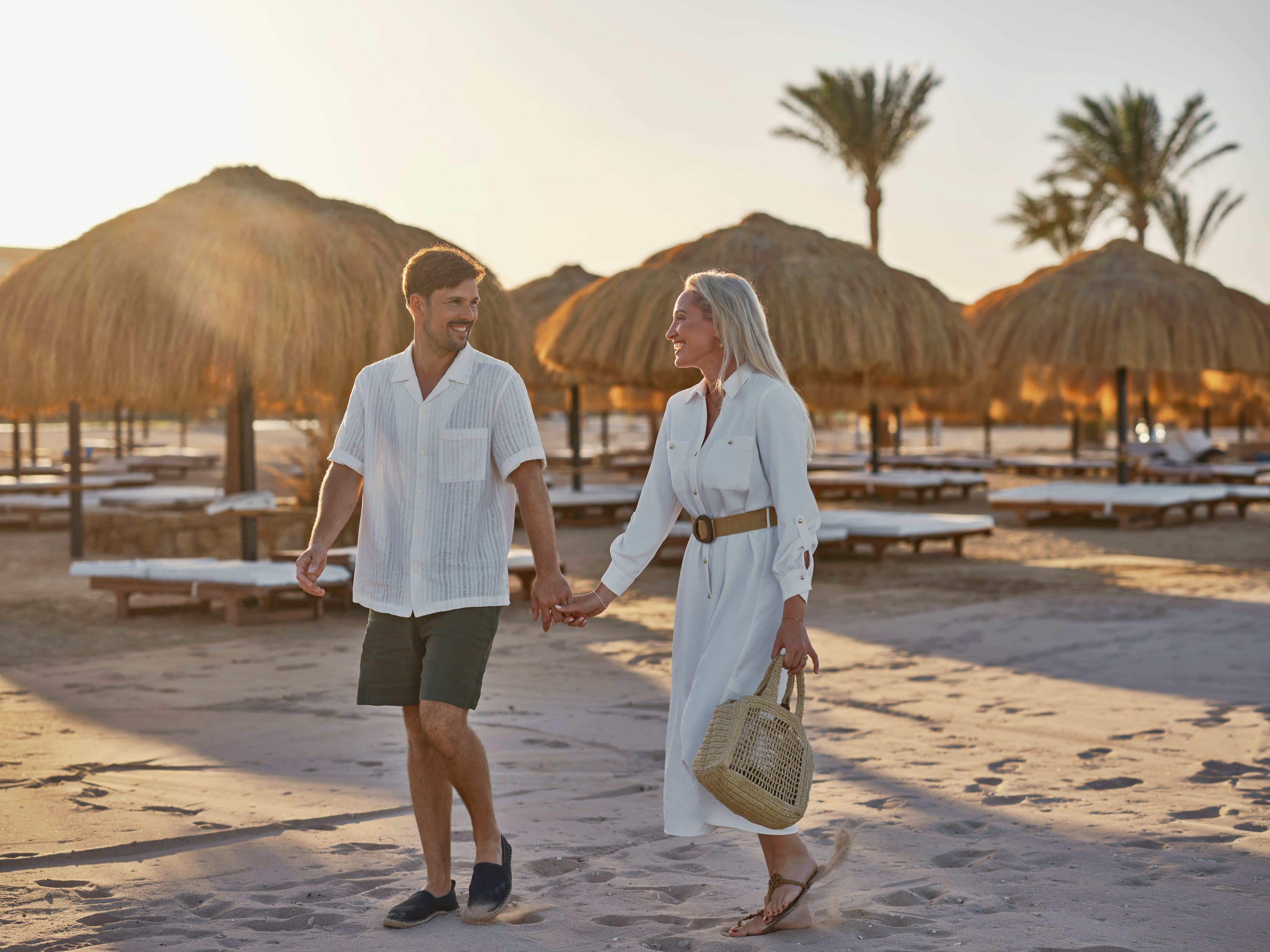 Paar spaziert am Strand bei Sonnenuntergang, mit Hotels im Hintergrund, entspannter Urlaub