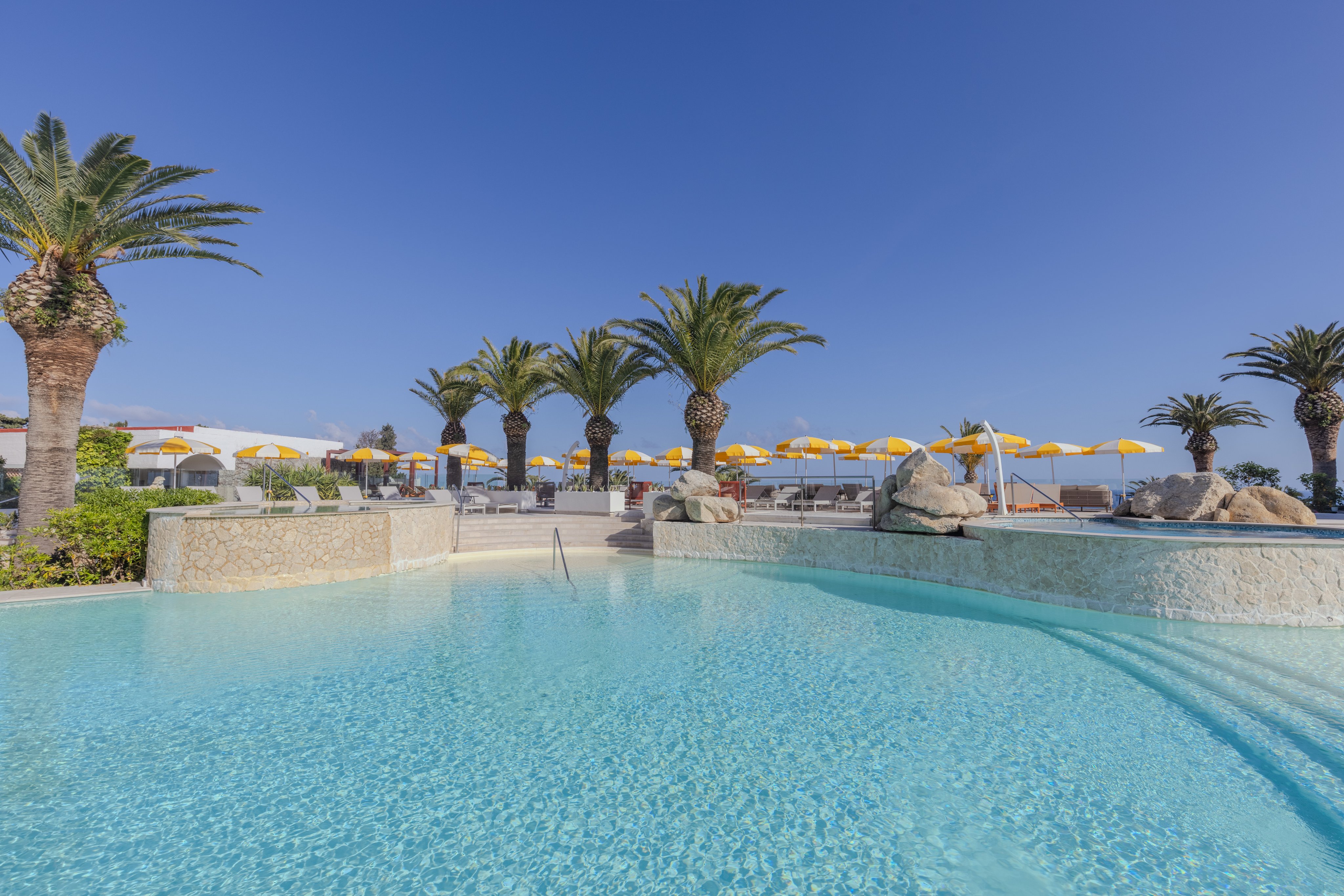 Blick auf einen Pool mit Palmen und Sonnenschirmen am Strand bei Aldiana Premium Cluburlaub