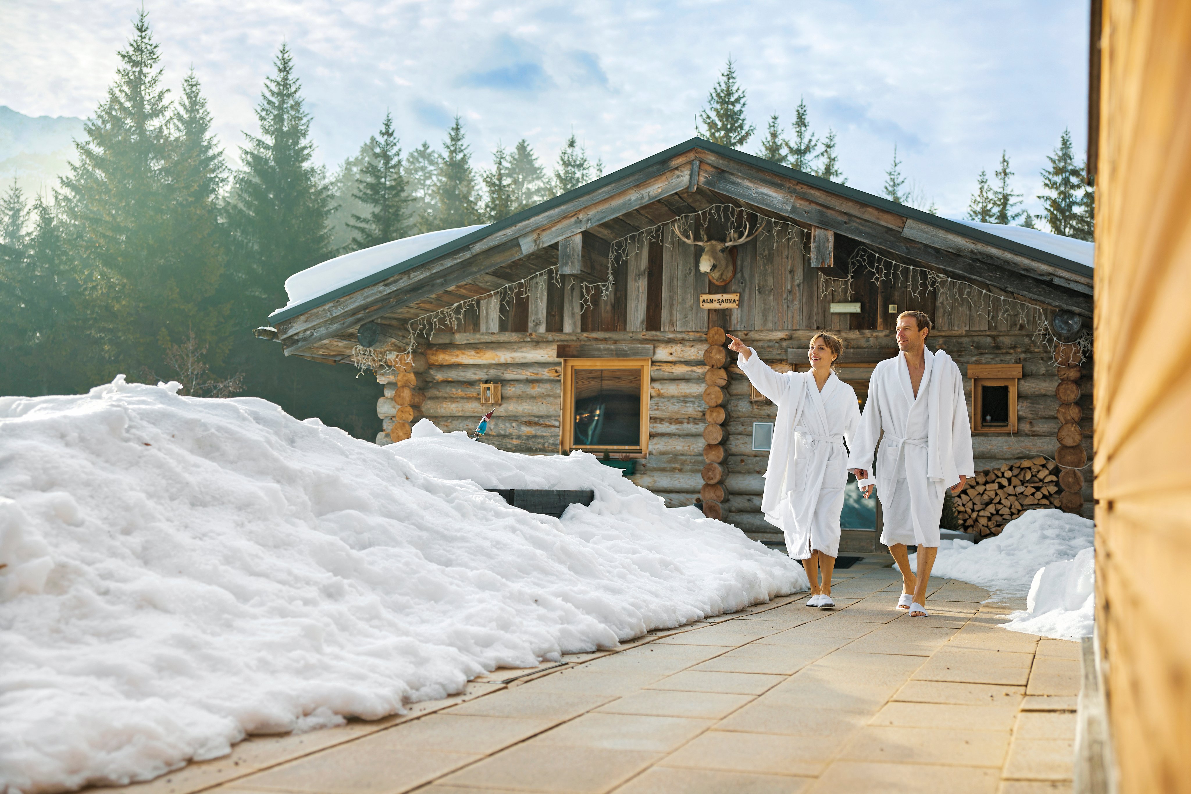 Paare im Bademantel vor einer Holzblockhütte im Schnee bei Aldiana Premium Cluburlaub