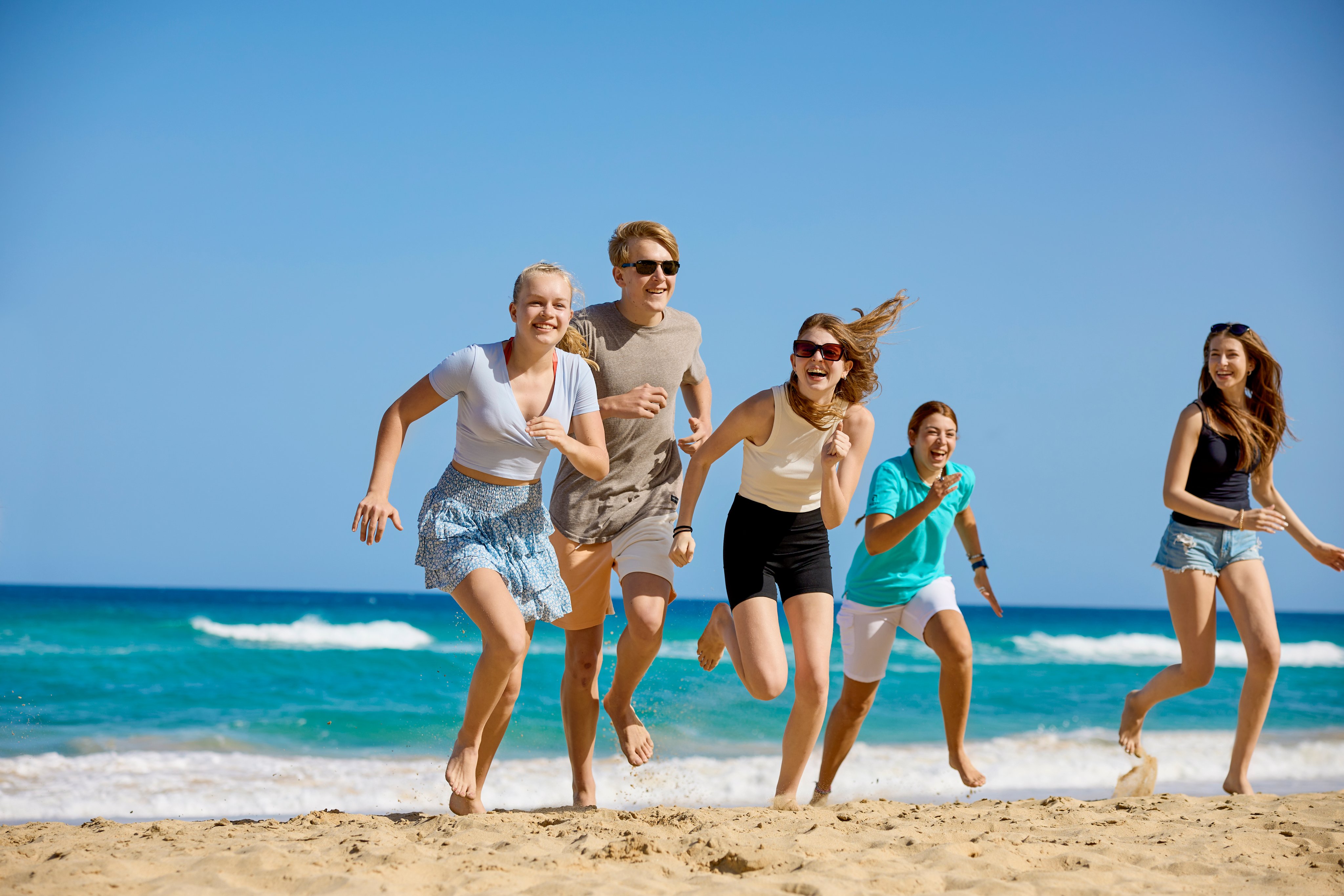 Familien beim Laufen am Strand bei Sonnenlicht, im Hintergrund das Meer und blauer Himmel