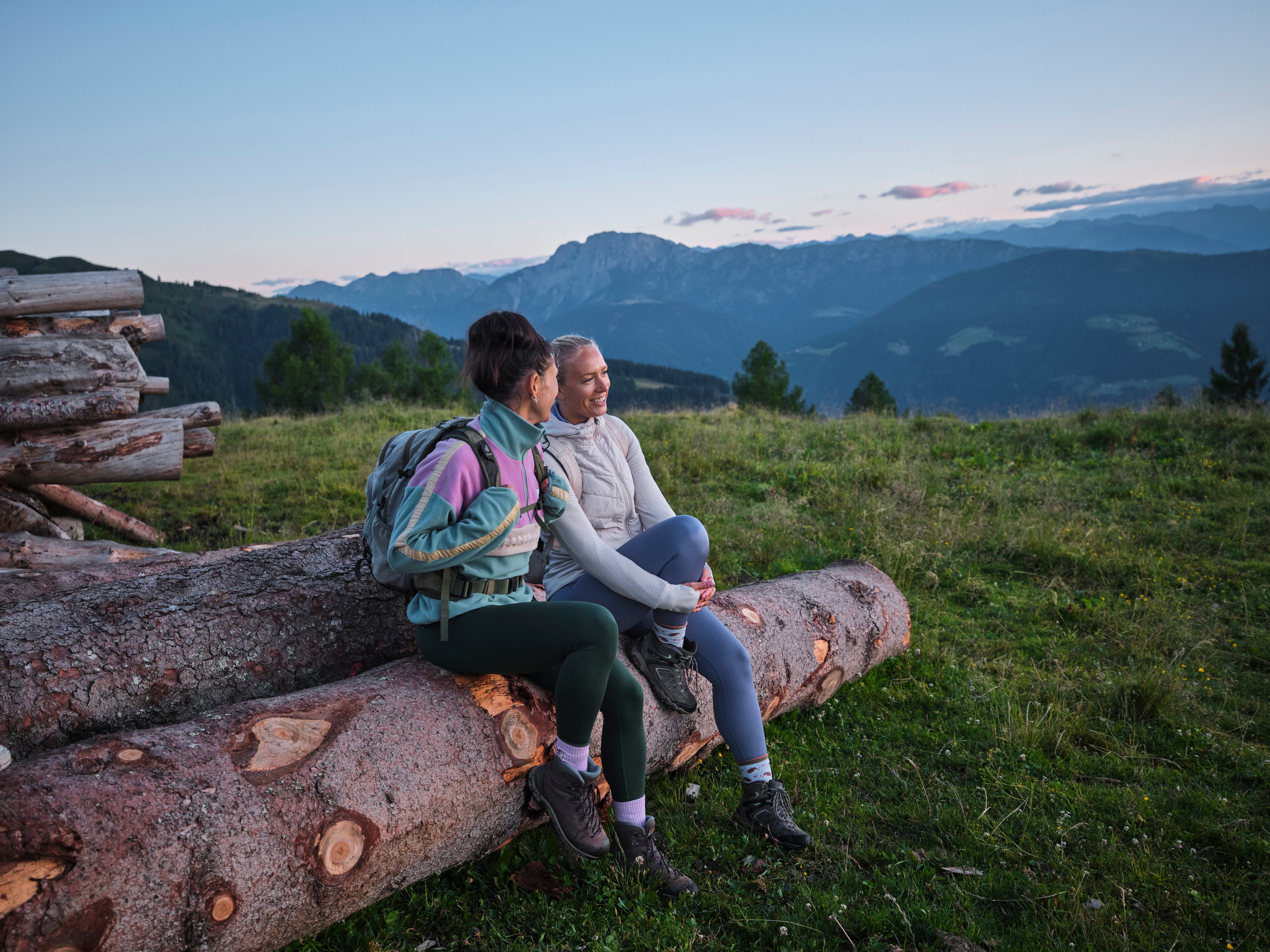 Zwei Frauen sitzen auf einem Baumstamm in der Natur mit Bergblick, Entspannung bei Aldiana.