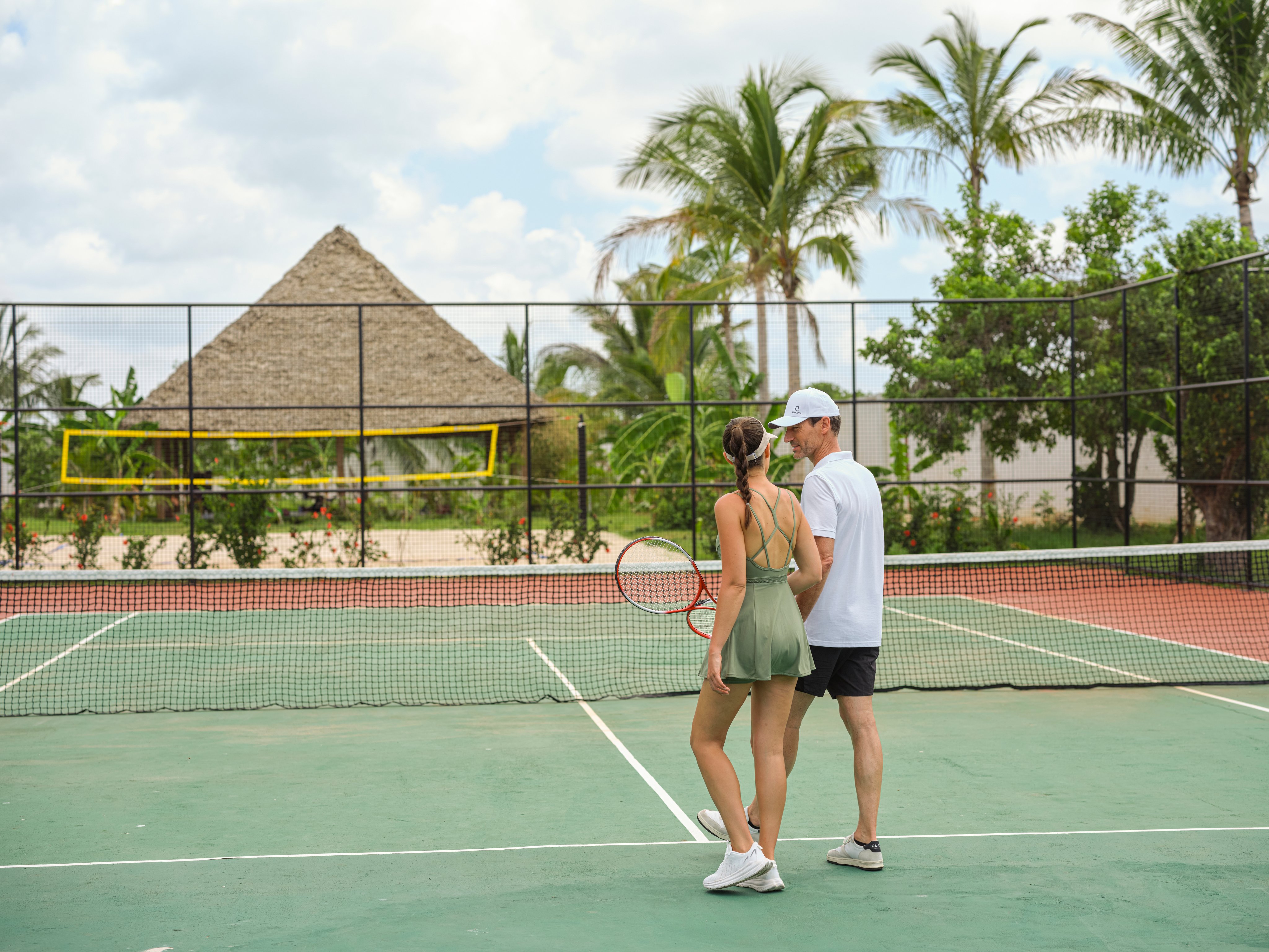Paar spielt Tennis auf einem Außengelände mit Palmen und Hütte im Hintergrund