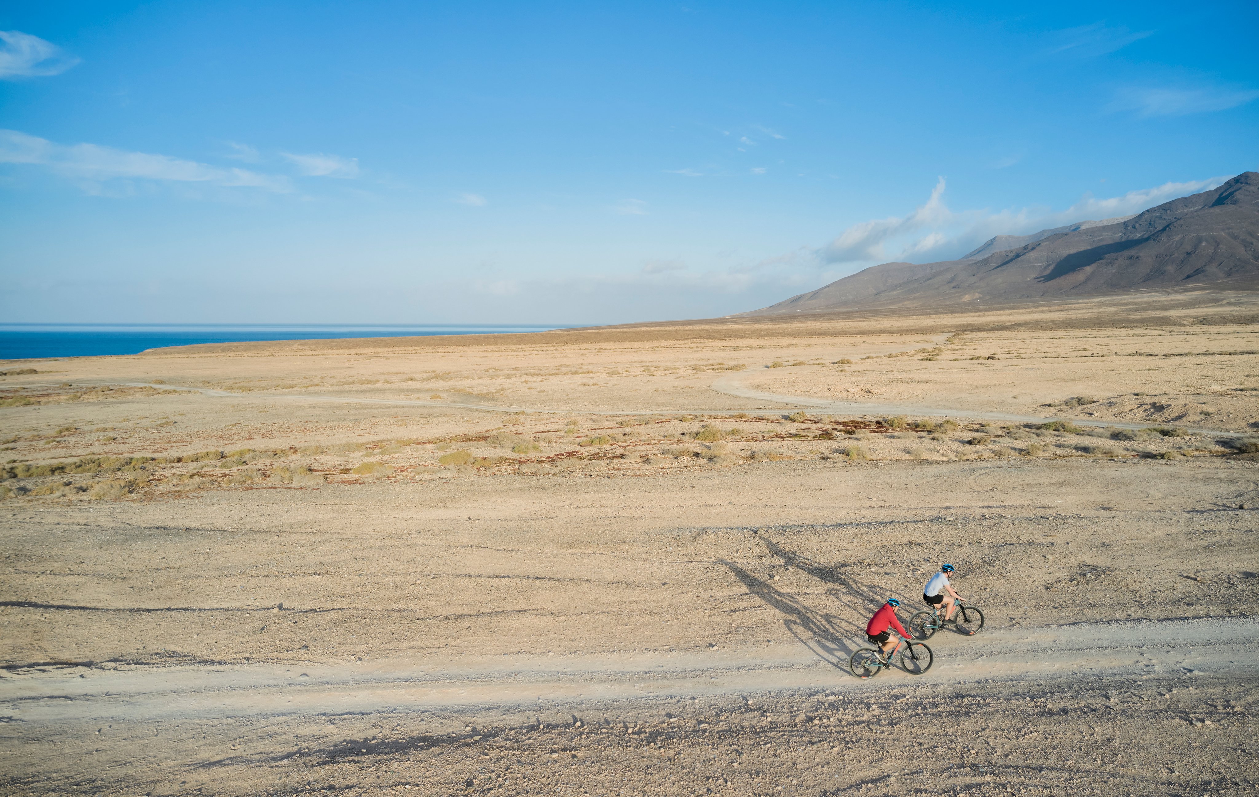 Zwei Radfahrer in einer weiten, kargen Landschaft mit Bergen im Hintergrund, bei Sonnenlicht.