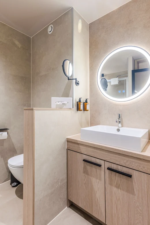 Ein modernes Badezimmer mit Waschtisch, rundem Spiegel mit LED Beleuchtung, WC und beige Fliesen