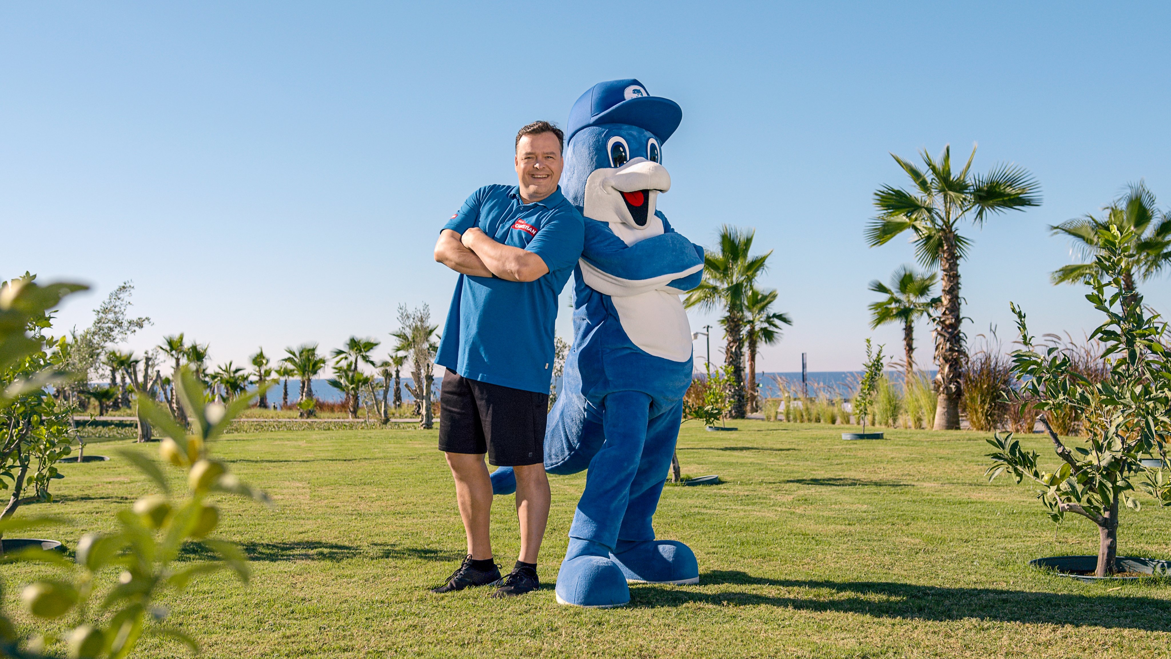 Freundlicher Mensch mit Sonnenbrille steht neben Counterpart im blauen Tierkostüm im grünen Park