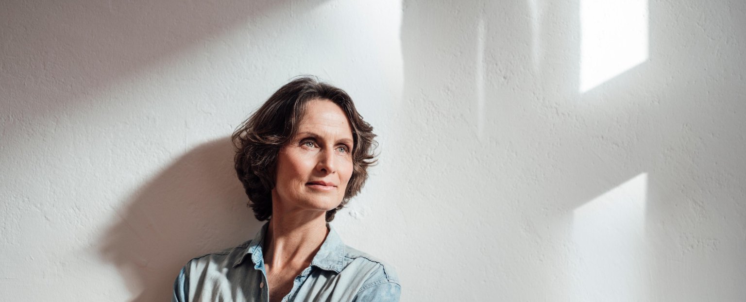 Eine nachdenkliche Frau mit lockigem Haar, die an einer weissen Wand lehnt, beleuchtet von sanftem Licht.