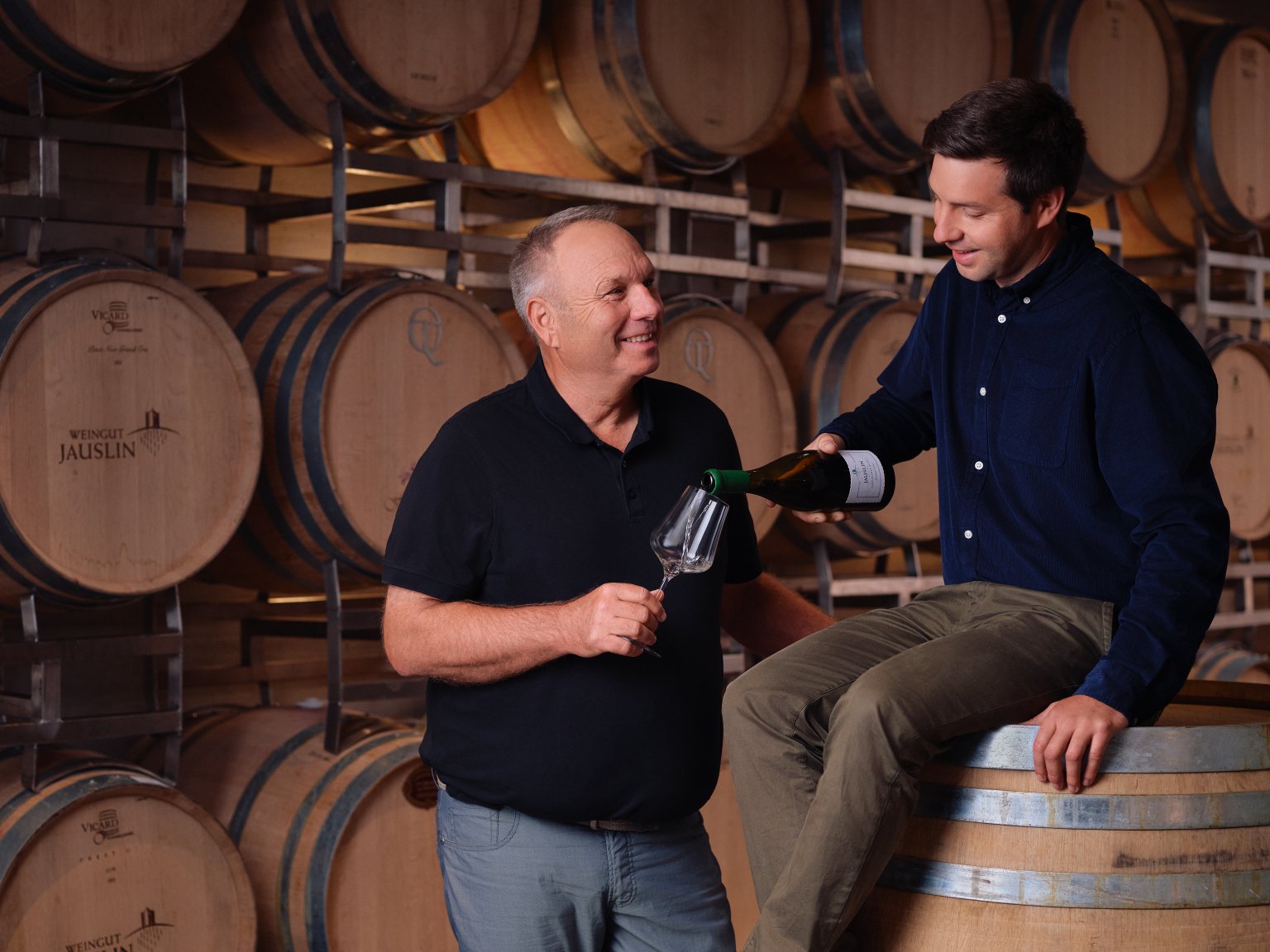 Zwei Männer im Weinkeller, einer schenkt dem anderen Wein ein.