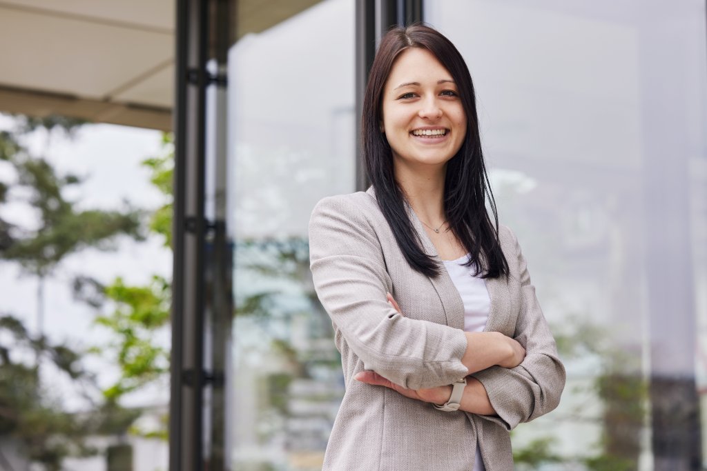 Frau im Anzug lächelt vor einem modernen Gebäude inmitten der Natur.