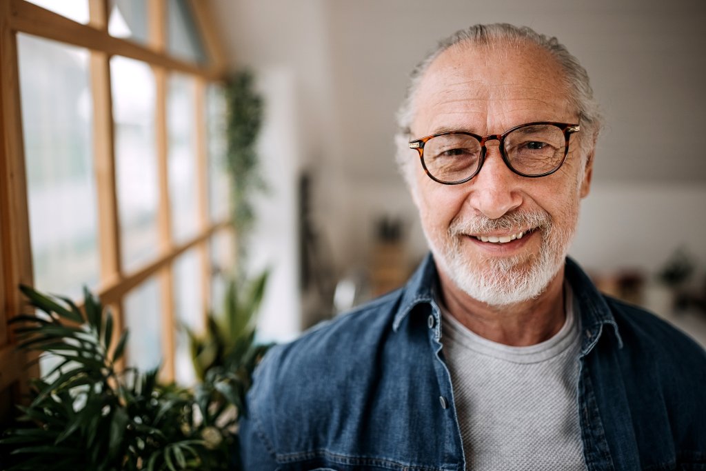 A smiling man with glasses and gray hair in a room with large windows.