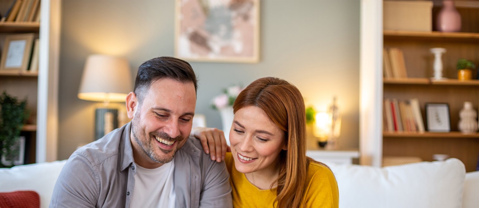 Zwei Personen sitzen auf einer Couch, schauen auf ein Smartphone, im Wohnzimmer.