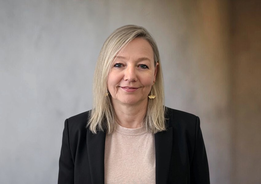 Frau mit blonden Haaren und schwarzem Blazer vor einfarbigem Hintergrund.