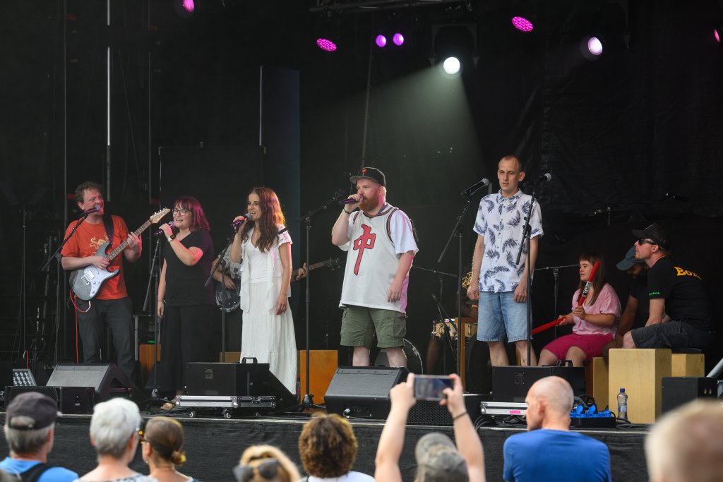 Sechs Menschen auf einer Bühne, fünf singen, einer spielt Gitarre, Publikum schaut zu