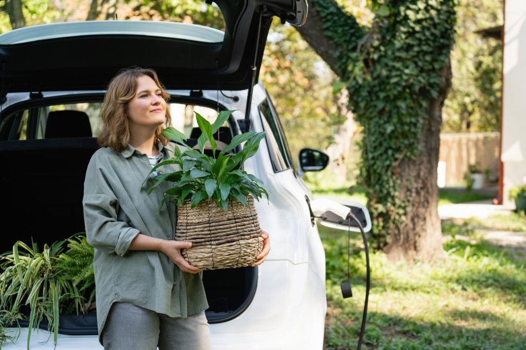 Frau mit Pflanze im Topf neben einem ladenden Elektroauto im Hof ​​eines Landhauses