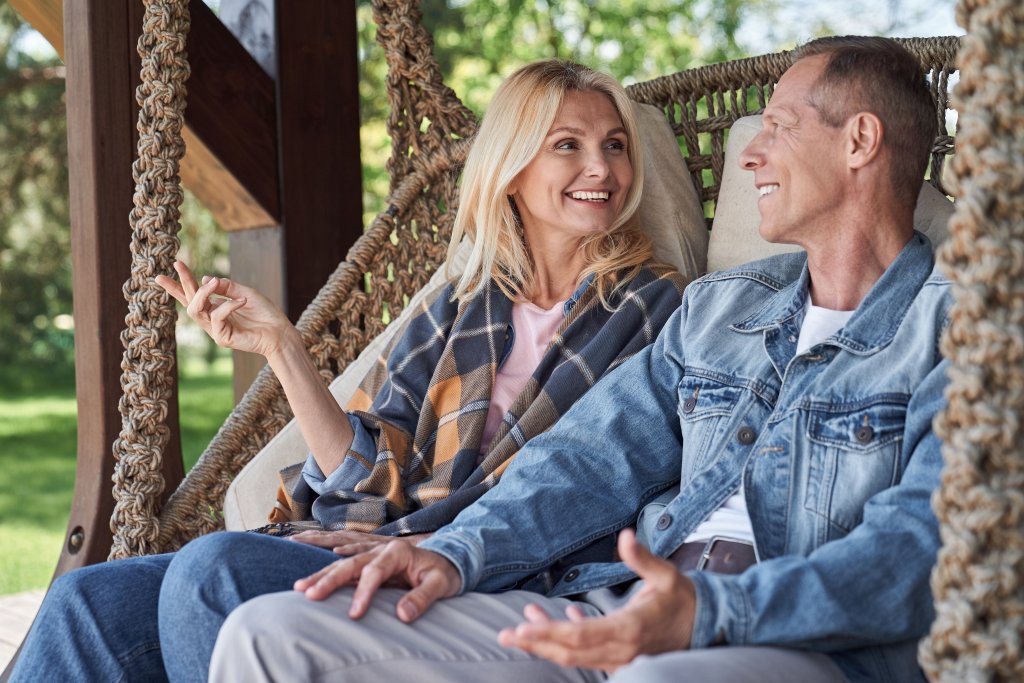 Zwei Menschen sitzen auf einer Schaukel im Garten, lachen und sprechen miteinander.