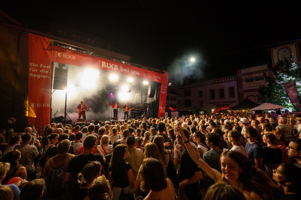 Ein Konzert bei Nacht mit einer großen Menschenmenge vor einer Bühne mit Rauch und Lichtern.