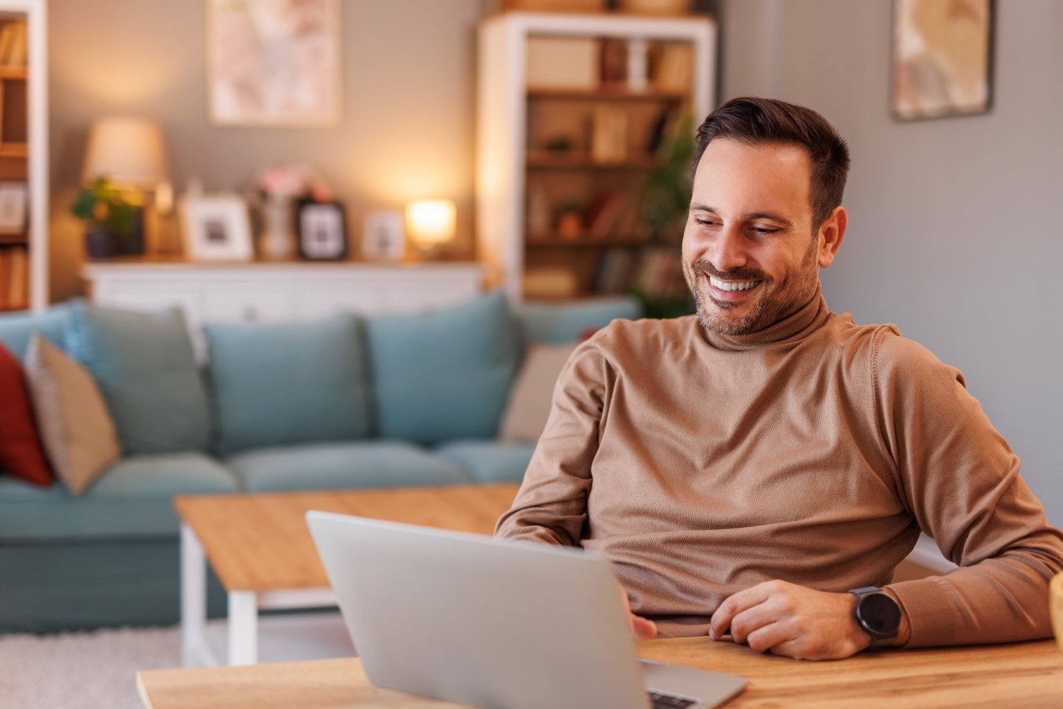 Mensch sitzt am Tisch mit Laptop vor einem gemütlichen Wohnzimmer im Hintergrund aus dem Artikel Der schnelle Gewinn oder langfristiger Vermögensaufbau?