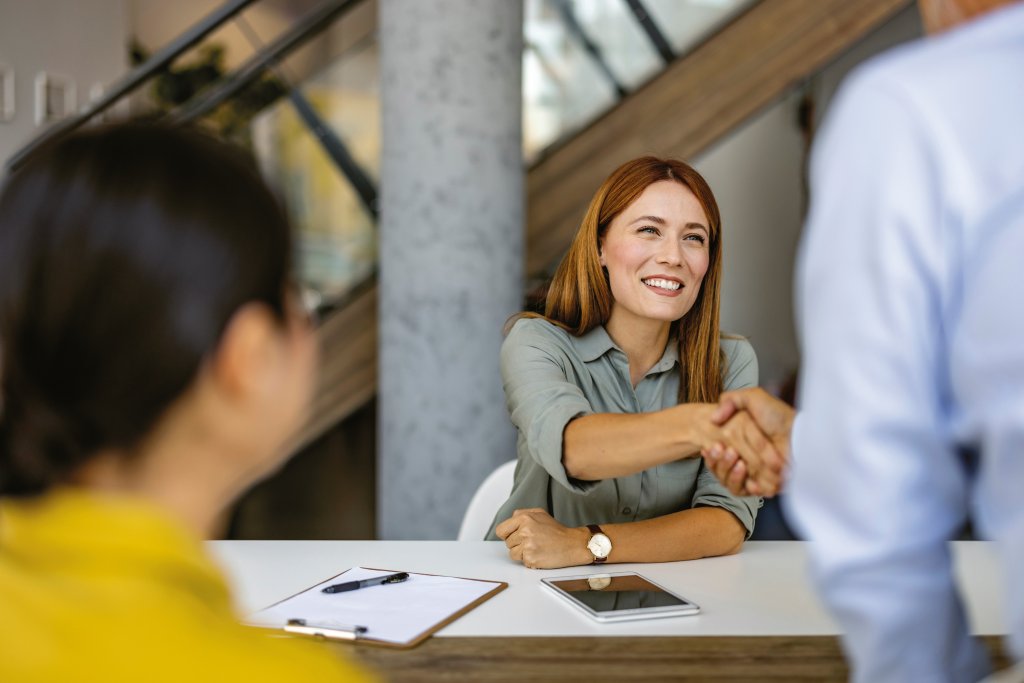 Eine lächelnde Geschäftsfrau begrüßt eine Kollegin per Handschlag über den Schreibtisch hinweg und demonstriert damit Professionalität und Teamgeist in einem modernen Büroumfeld.