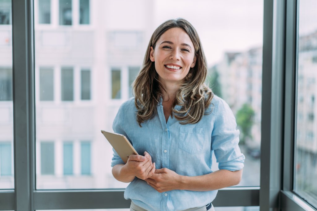Eine lächelnde Frau, die einen Ordner vor einem großen Fenster in einem modernen Büro hält.