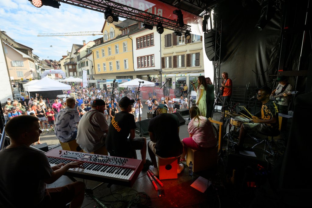 Menschen auf einer Bühne spielen Musik vor einer Menschenmenge in einer Stadt.