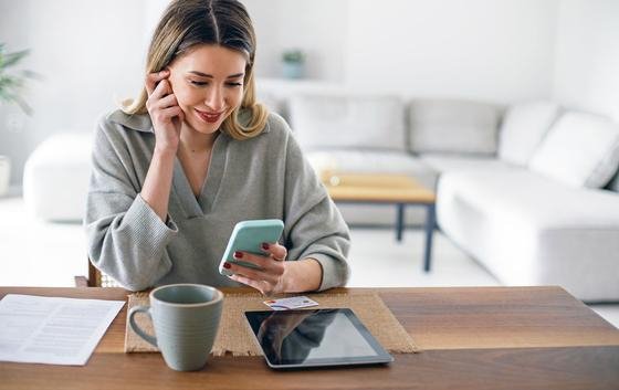 Eine junge Frau sitzt am Esstisch und bezahlt mit dem Smartphone ihre Rechnungen. Auf dem Tisch liegt ein Tablet und die Kreditkarte.