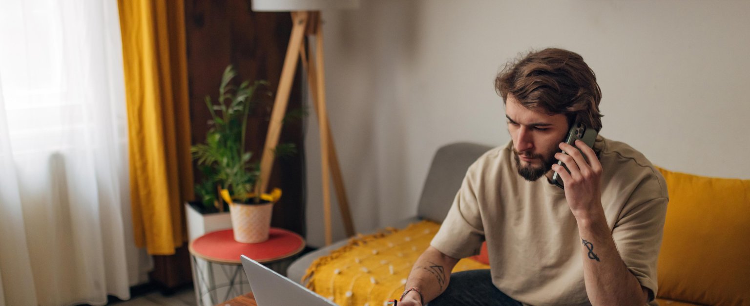 Mann, der am Telefon spricht, während er an einem Laptop in einem gemütlichen Raum mit Pflanzen und Dekoration arbeitet.