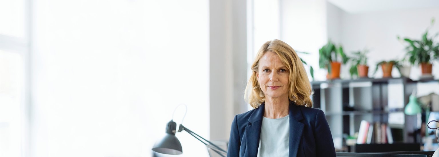 Frau steht im Büro vor einem Schreibtisch mit Lampen und Regalen im Hintergrund.