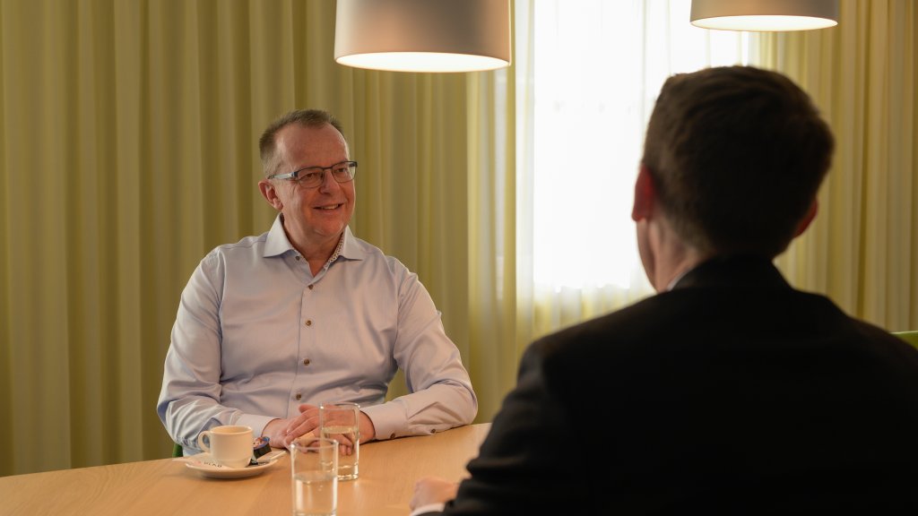 Zwei Männer im Gespräch bei einem Kaffee im Büro, im Hintergrund Vorhänge und Lichtkuppeln