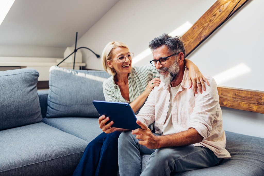 Ein älteres Paar sitzt auf einem gemütlichen Sofa und nutzt gemeinsam freudig ein Tablet in seinem modernen Zuhause. Warme, liebevolle Szene des Ruhestands.