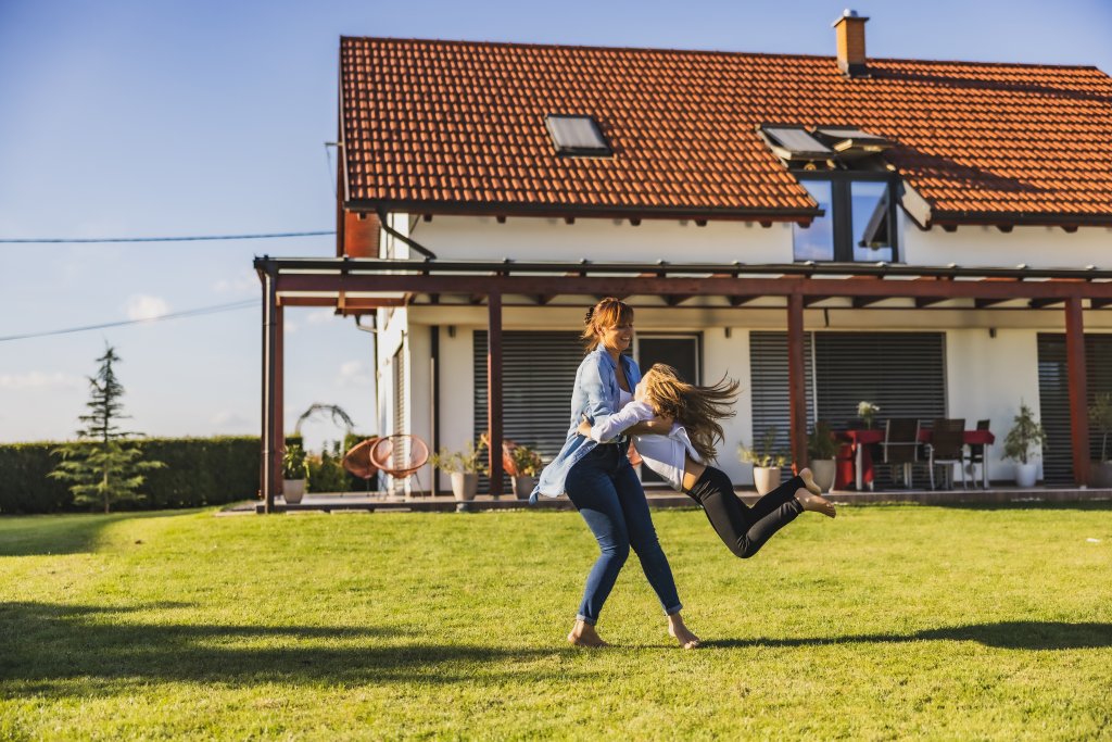 Zwei Frauen spielen lachend auf grünem Rasen vor einem Haus mit rotem Dach.