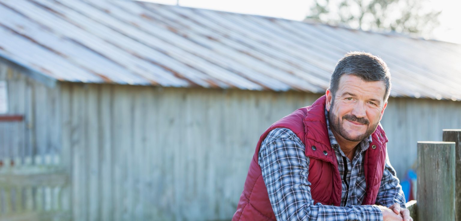 Mensch sitzt draussen an einem Zaun mit einer alten Scheune im Hintergrund